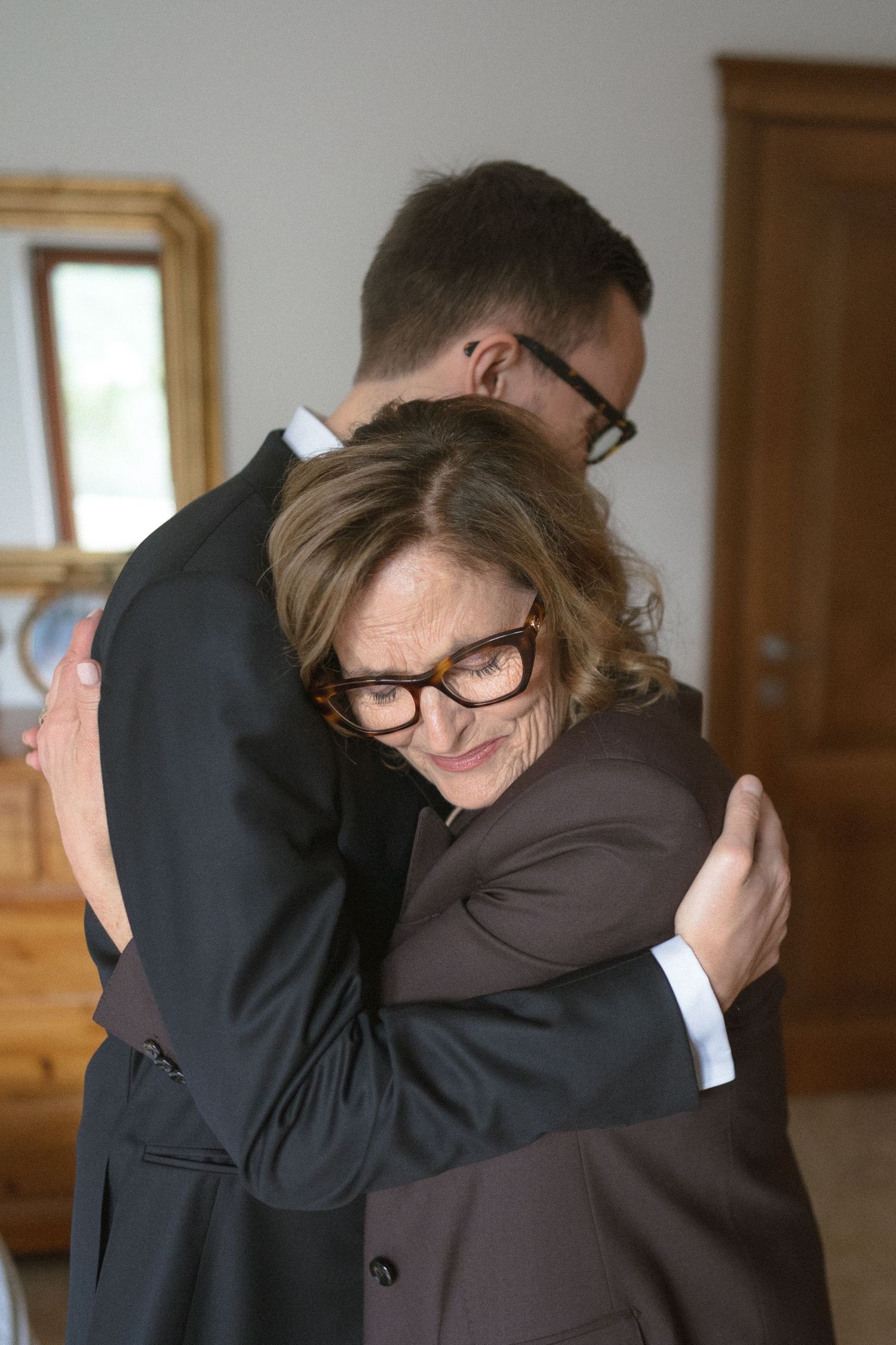 A woman in glasses smiles warmly while embracing a young man in a dark suit indoors.