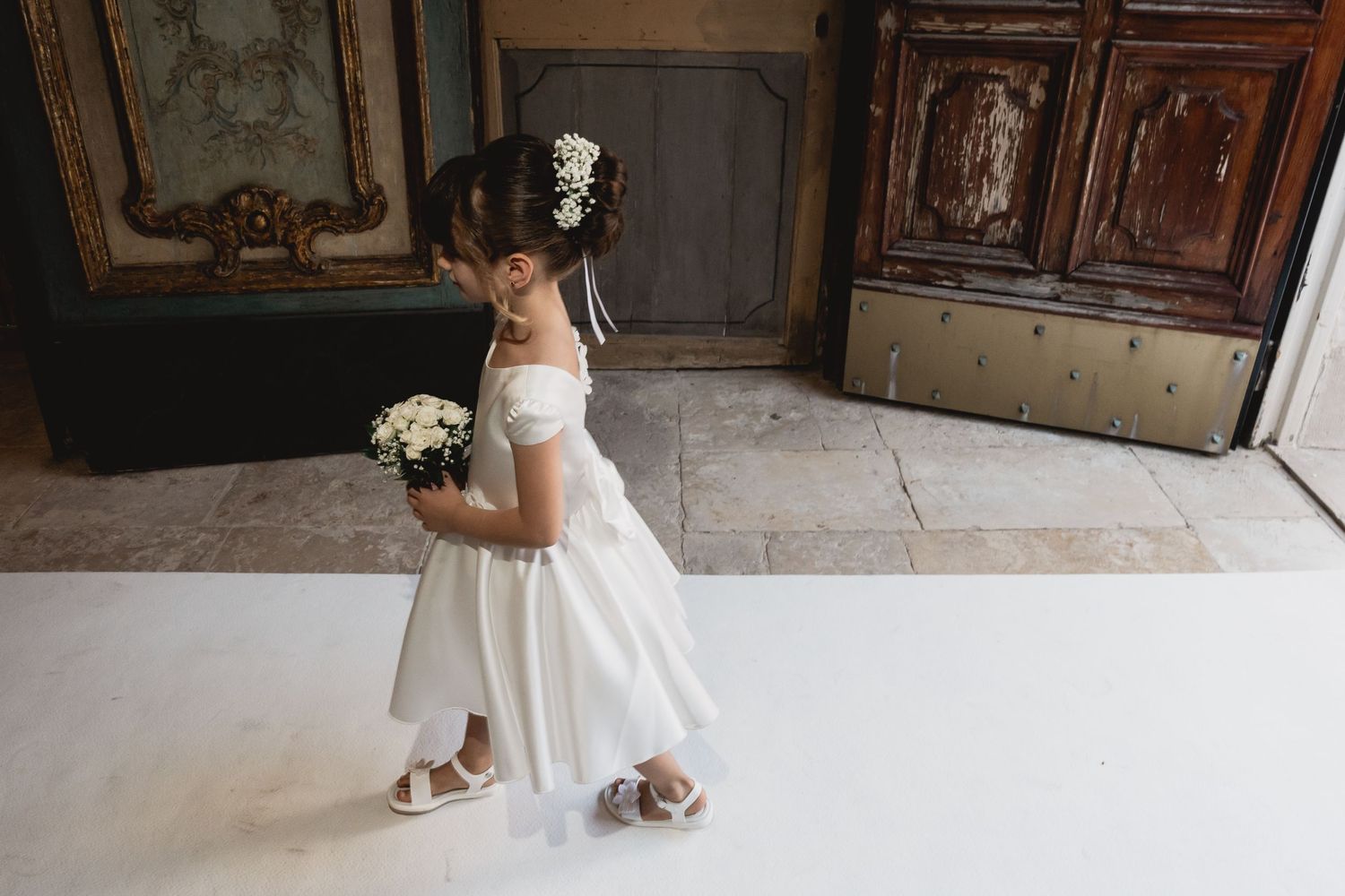 Young attendant in white dress holding bouquet stands in elegant interior setting.