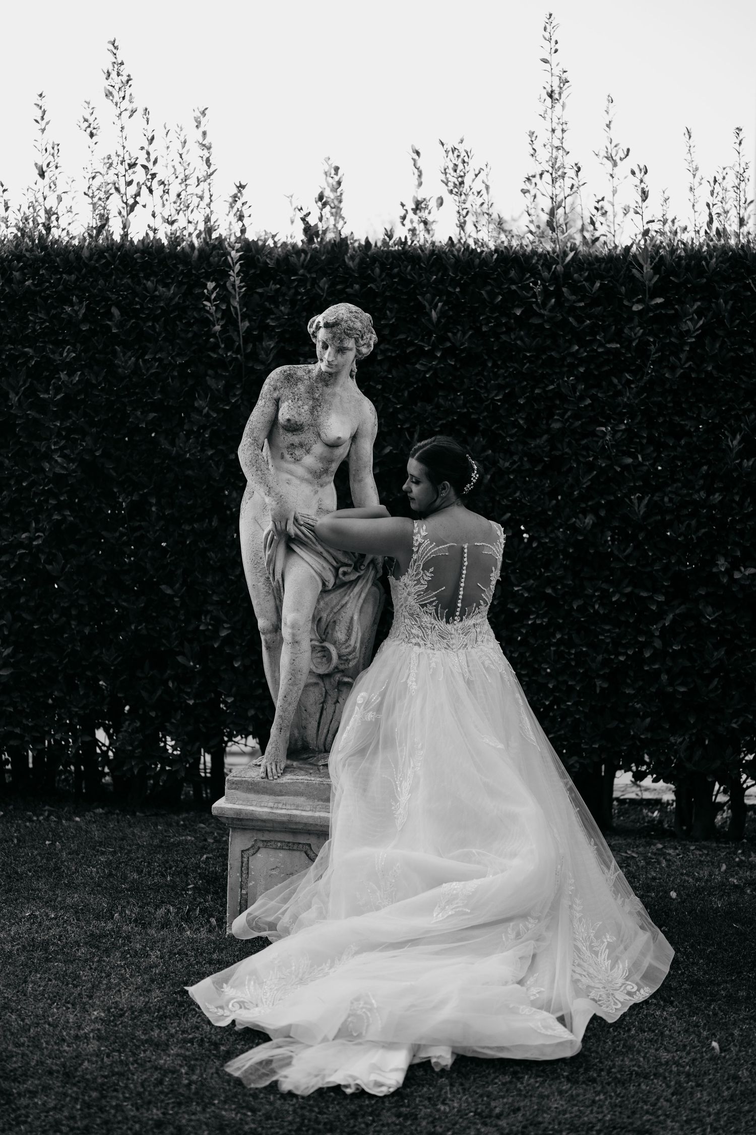 A bride in a flowing white gown poses next to a classical garden statue against a manicured hedge.