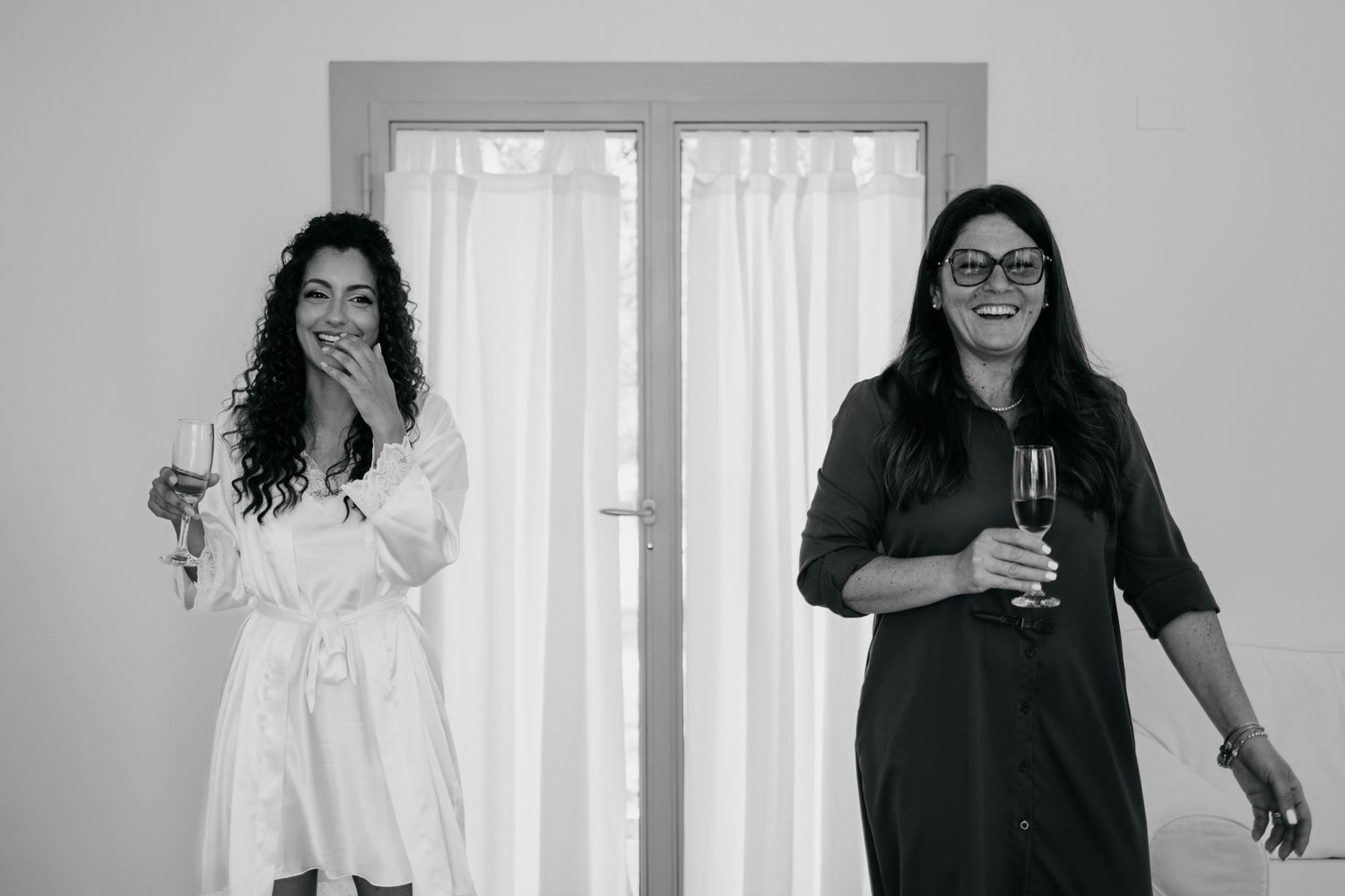 Two women laughing and holding drinks during wedding preparations.
