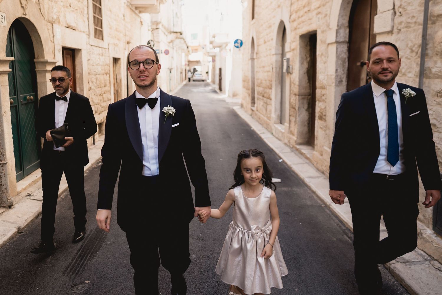 Wedding party walks down a narrow European stone street on a sunny day.