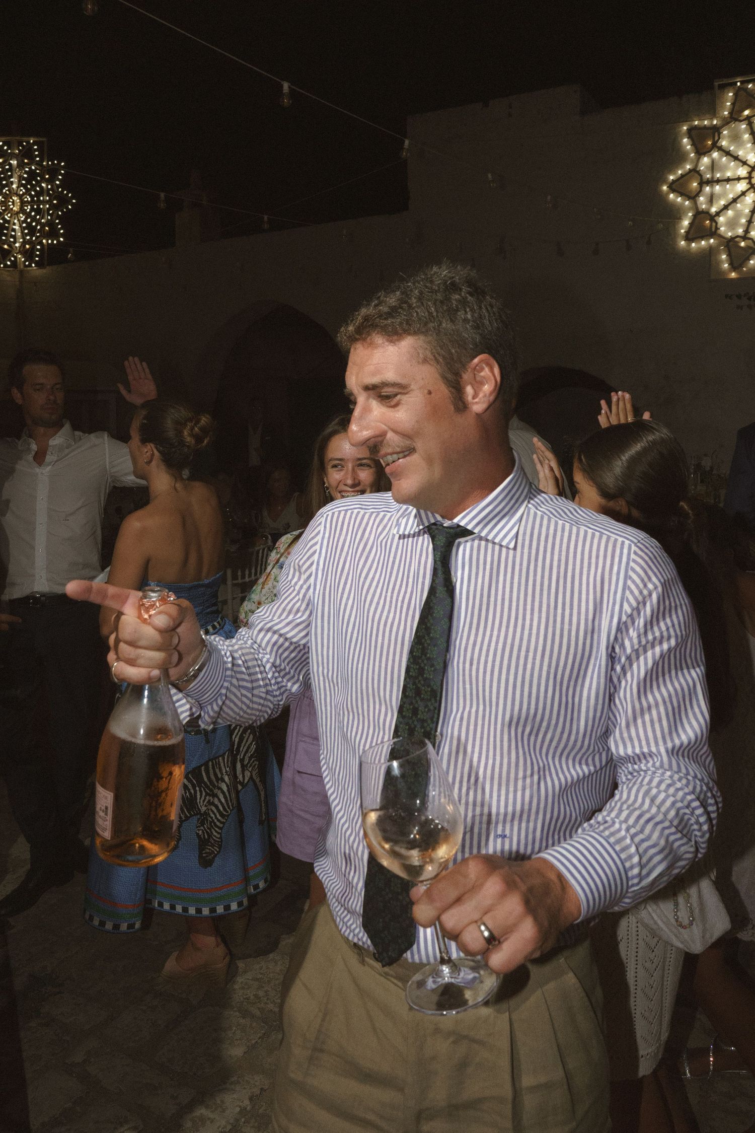 A person in a striped shirt and tie holds drinks while celebrating at a party with festive lights in the background.