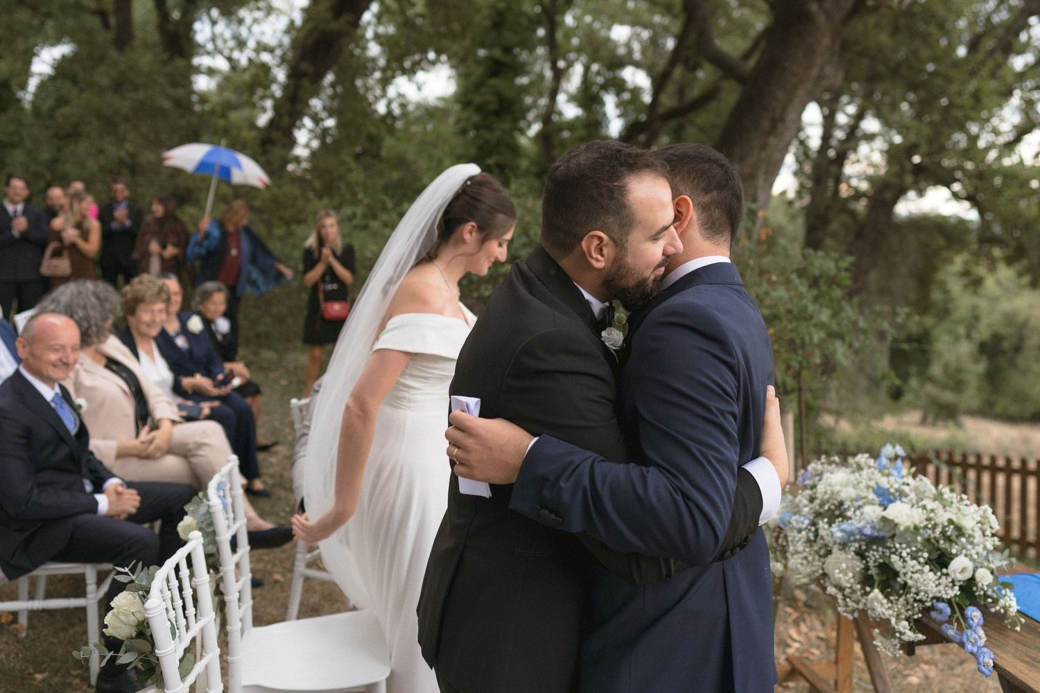Romantic wedding moment under trees as newlyweds share their first kiss while guests applaud.