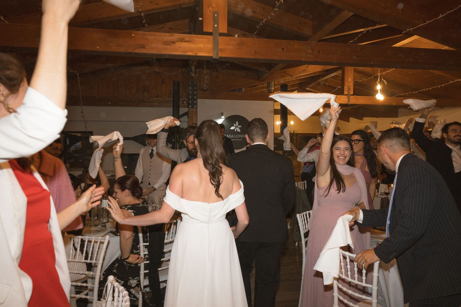 Wedding guests dancing and celebrating in a rustic venue with wooden beams and string lights overhead.