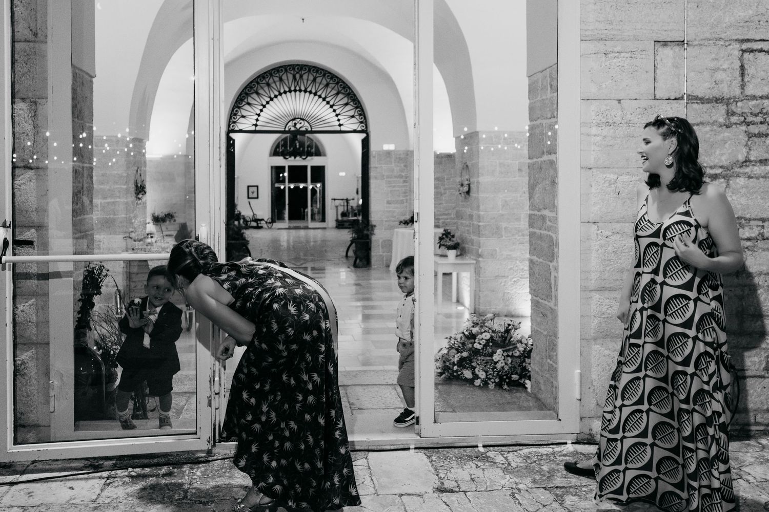 Black and white photograph of people in patterned dresses standing near arched entrance of a building.