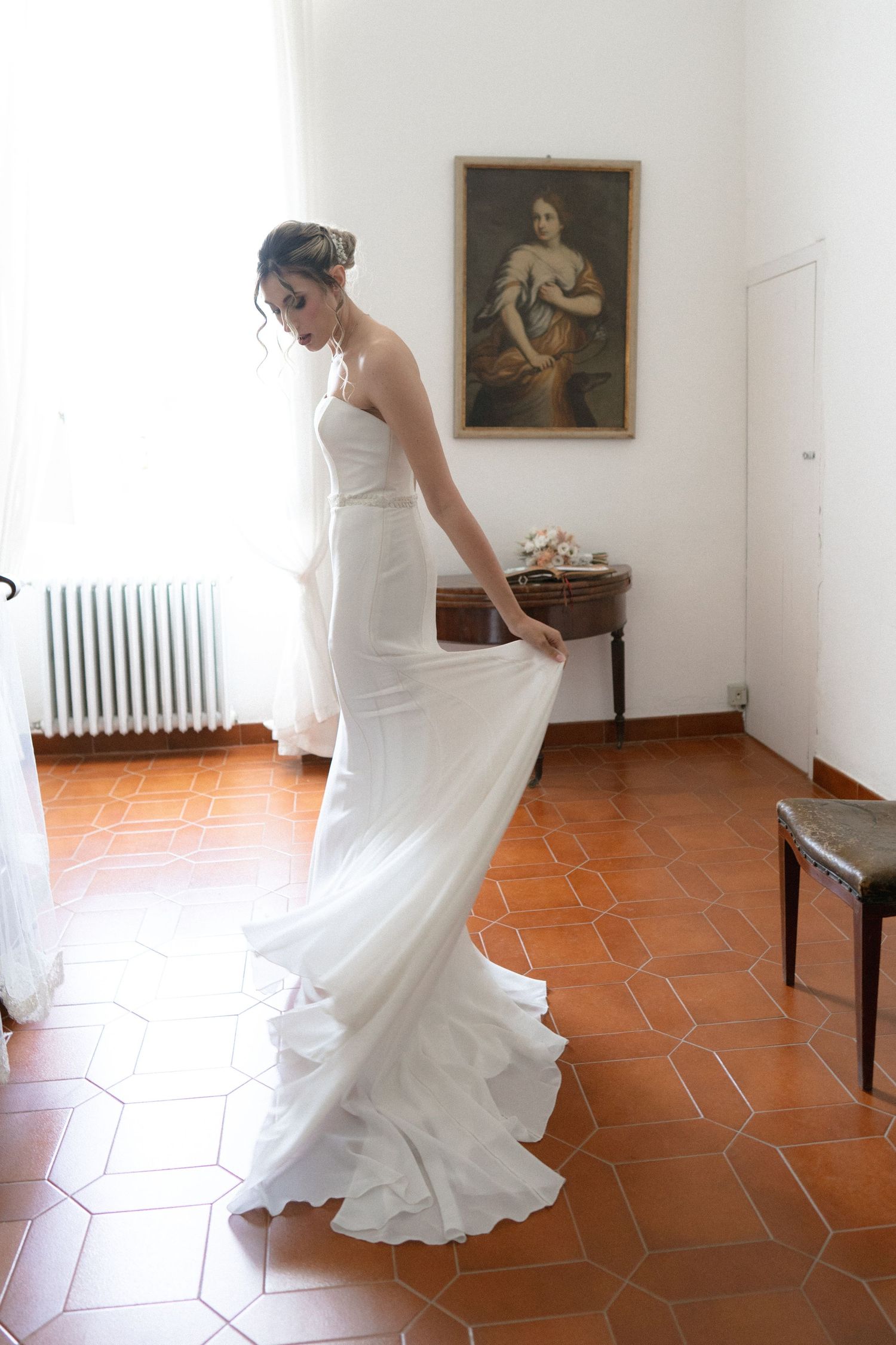 A white wedding dress twirls gracefully on terracotta tiles in a room with vintage paintings on the wall.
