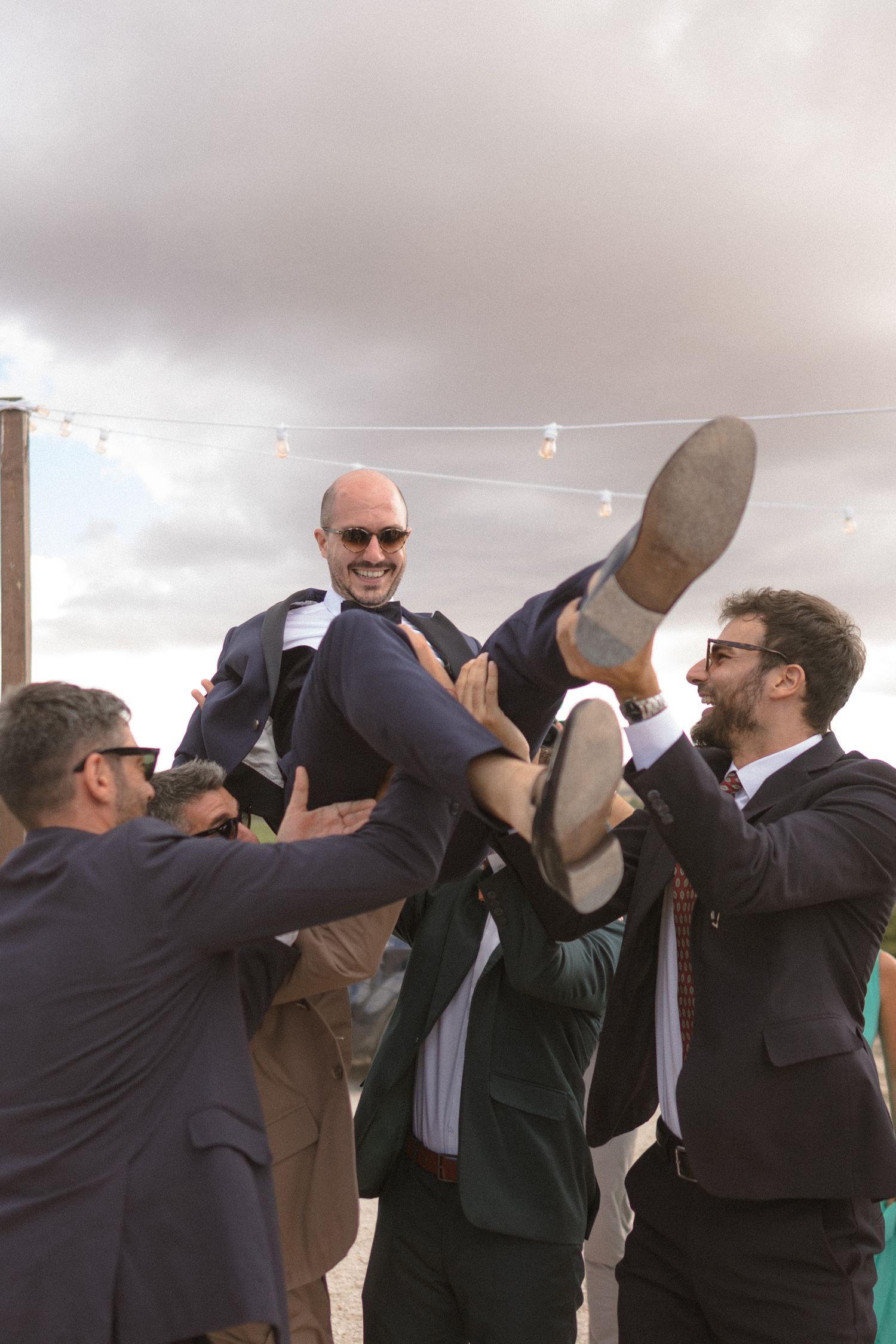 Group of groomsmen in suits performing a synchronized boot shot drinking ritual at an outdoor event.