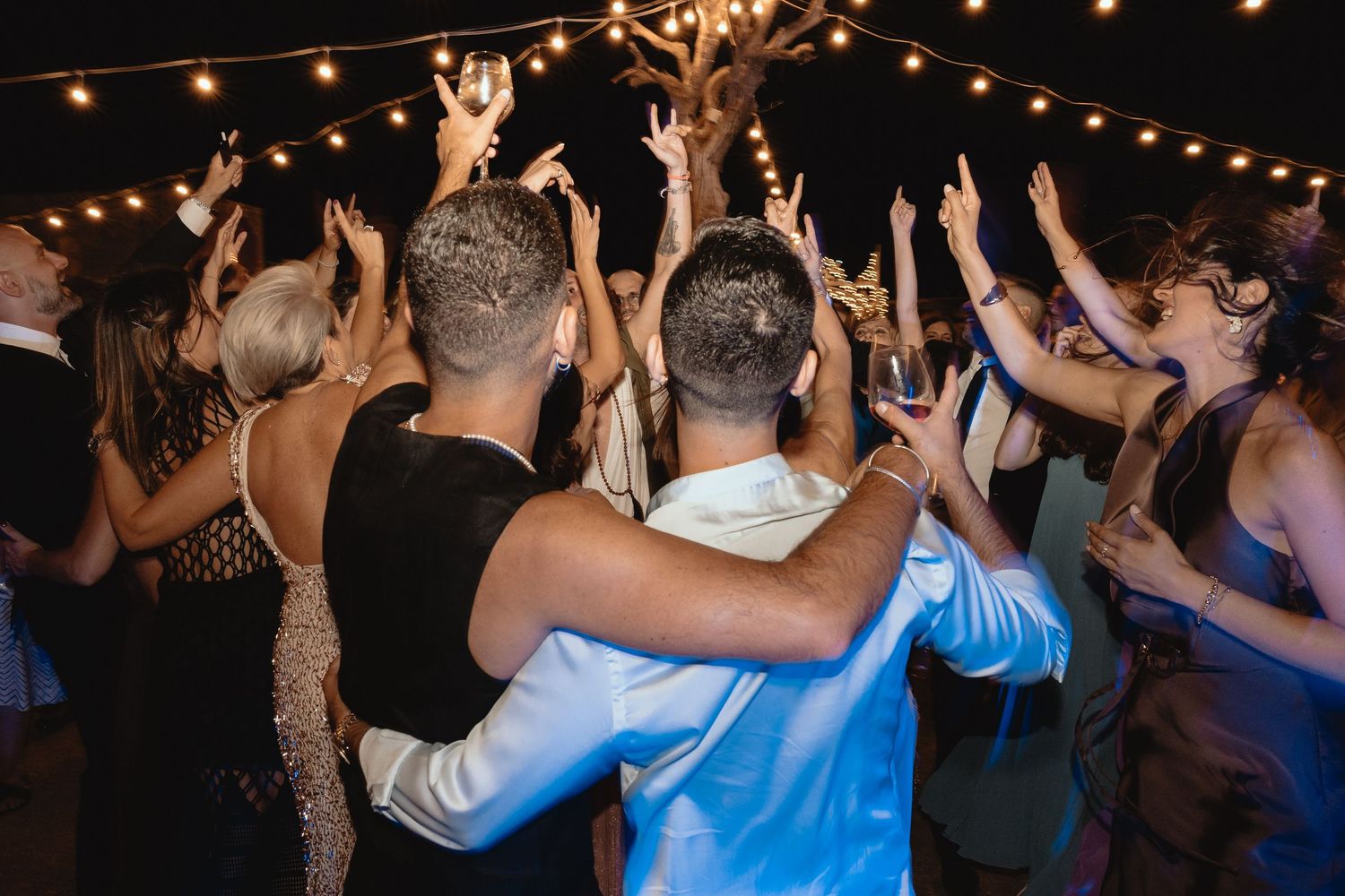 Wedding guests dancing under string lights at an outdoor evening reception.