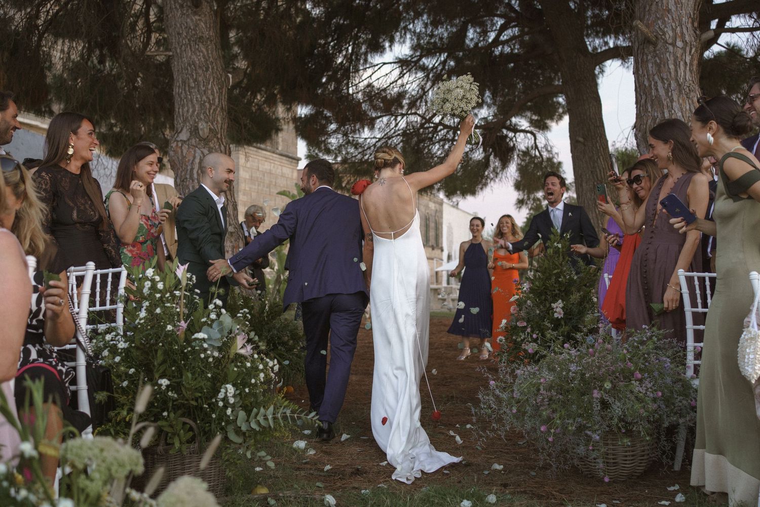 Wedding celebration on a garden lawn with guests gathered around white chairs and greenery.