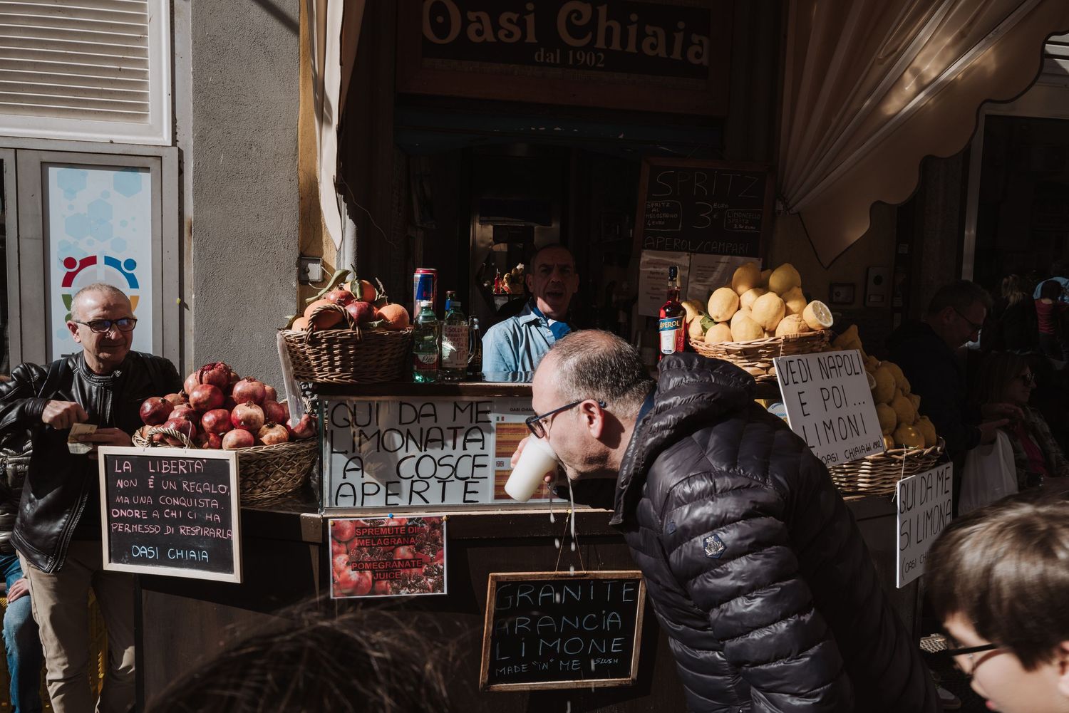 Shoppers browse fresh produce and goods at an Italian market stall with chalkboard signs displaying prices.