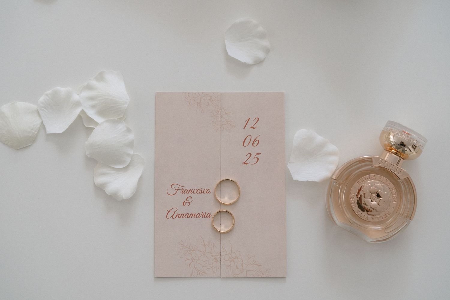 Wedding rings and delicate white rose petals arranged on a light pink invitation card.