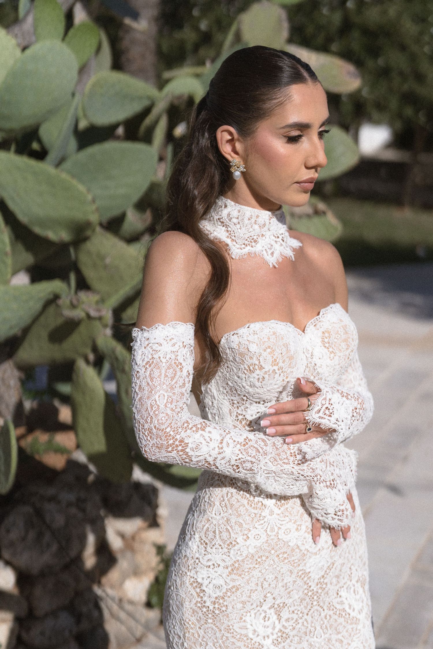 Bride in an off-shoulder white lace wedding gown with a high lace collar standing beside large prickly pear cactus leaves.