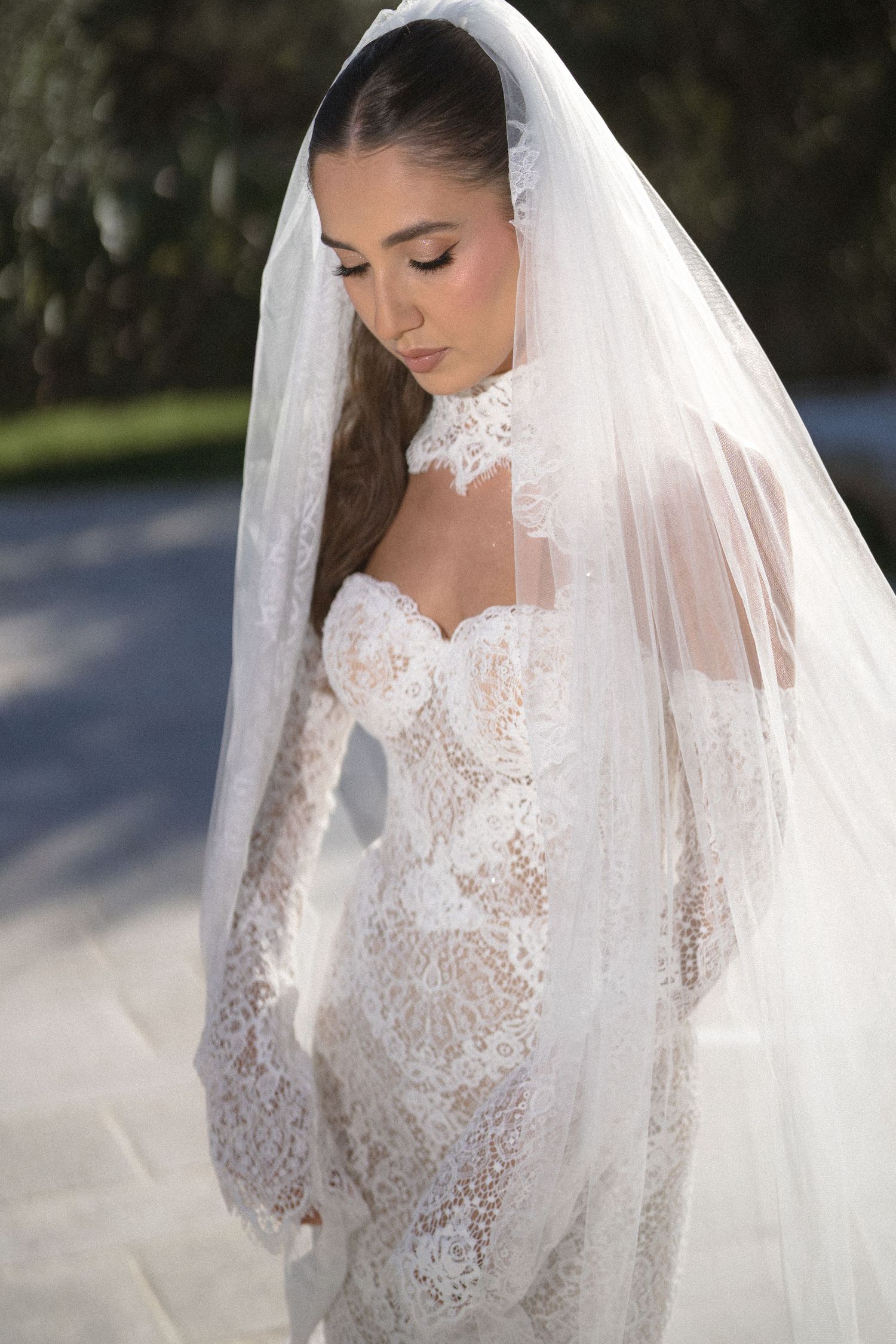 Bride in a strapless lace mermaid gown with a high lace collar and long sheer veil looking down outdoors.