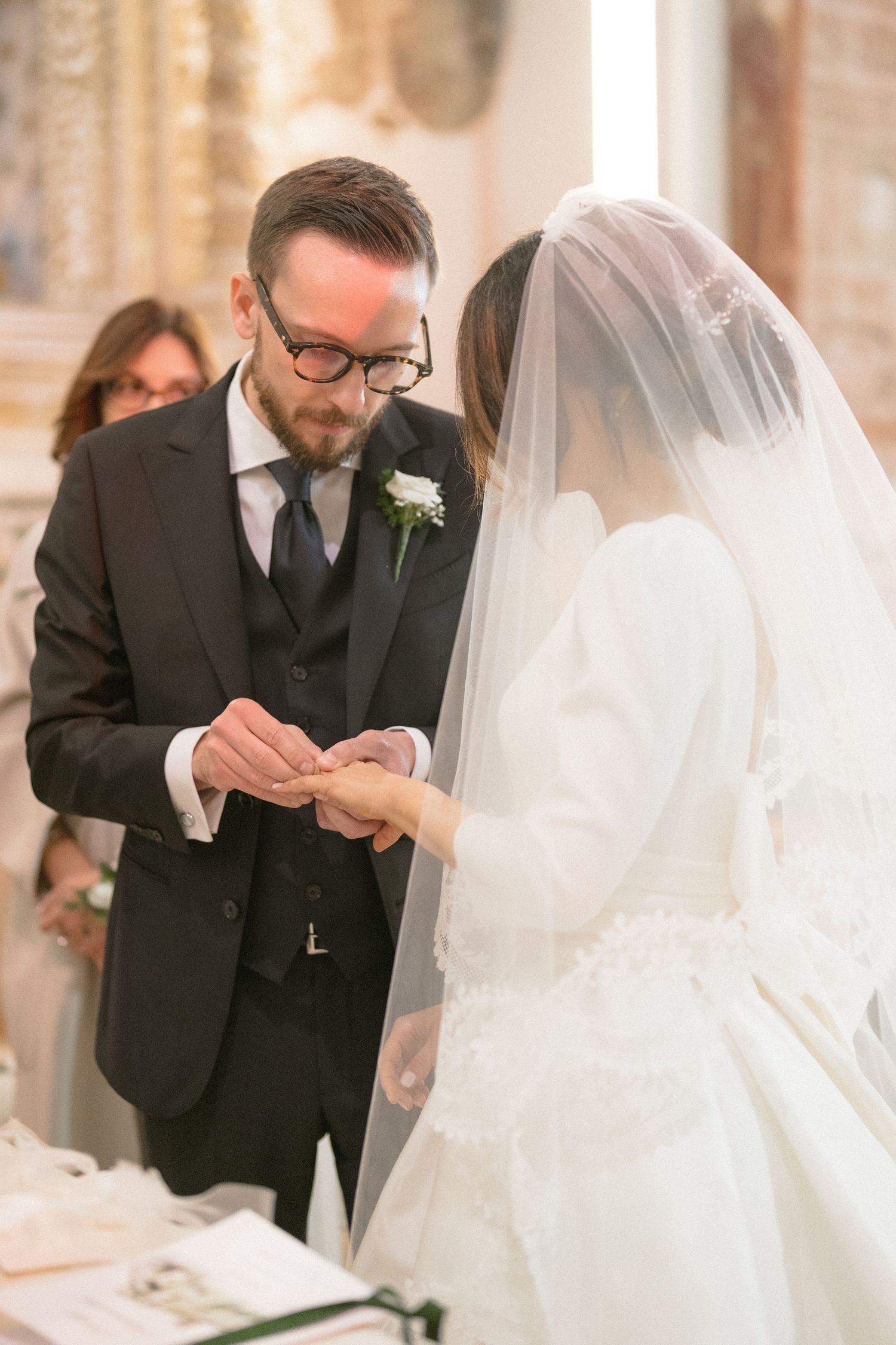 Groom with glasses places wedding ring on brides finger during church ceremony exchange of vows.