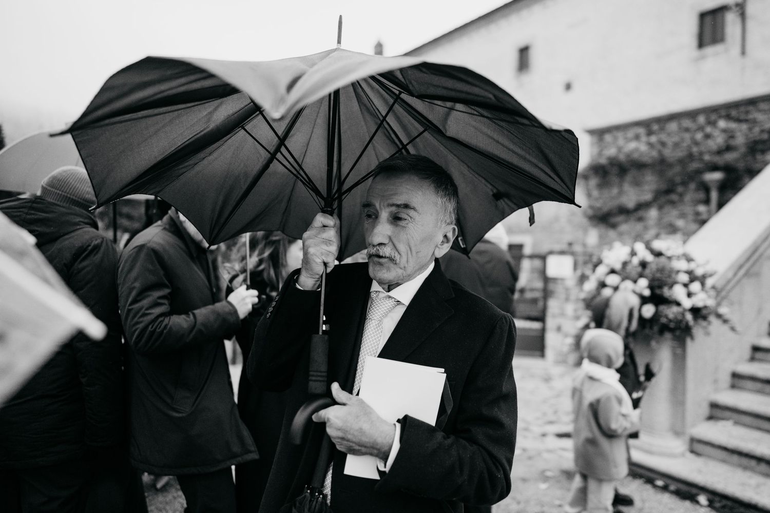 Elderly man in suit holds black umbrella outside rainy wedding venue in black and white photo.