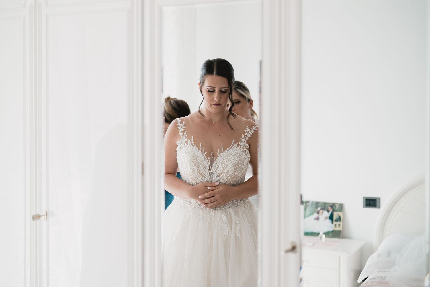 Bride in a white lace wedding dress stands in front of a doorway during wedding preparations.