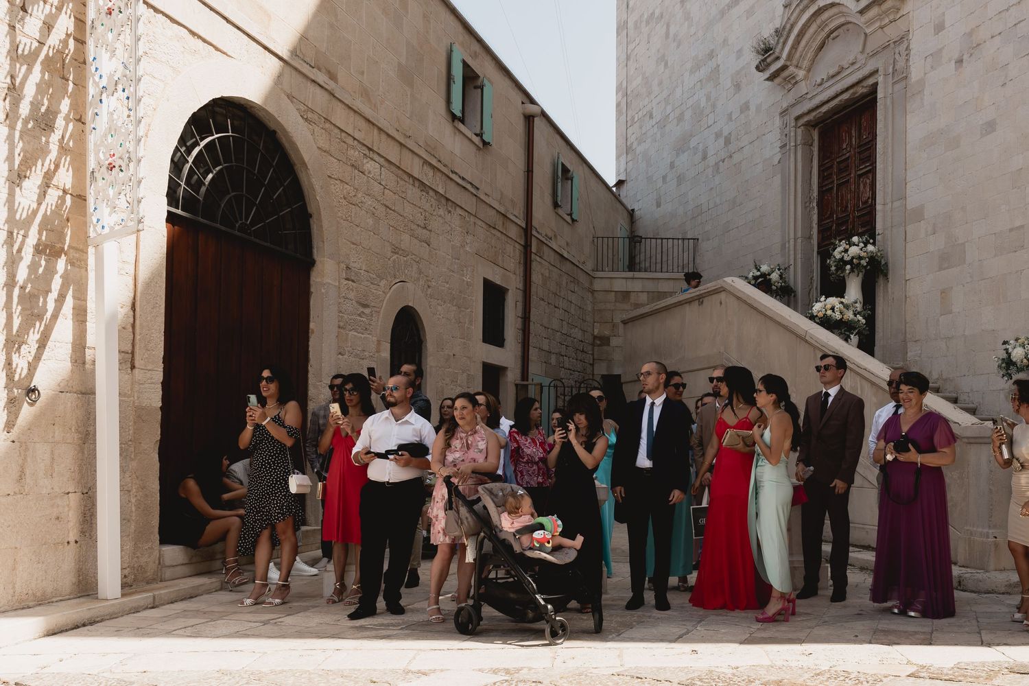 Wedding guests gather outside a stone building entrance in formal attire.