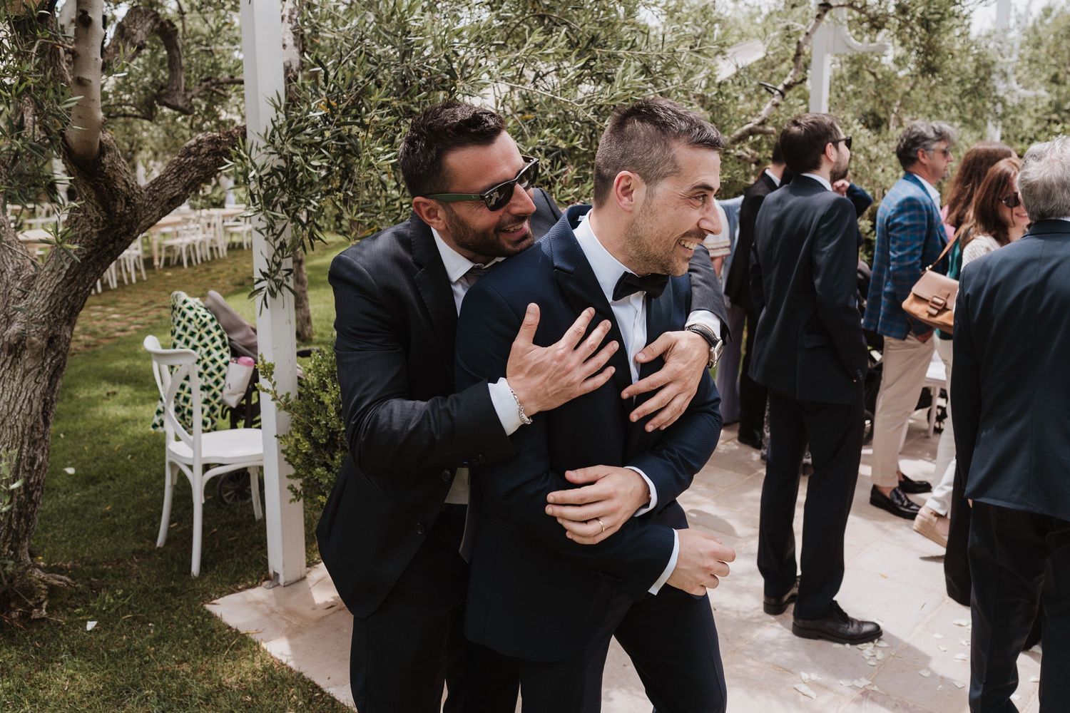 Two men in dark suits share an emotional embrace at an outdoor wedding reception.
