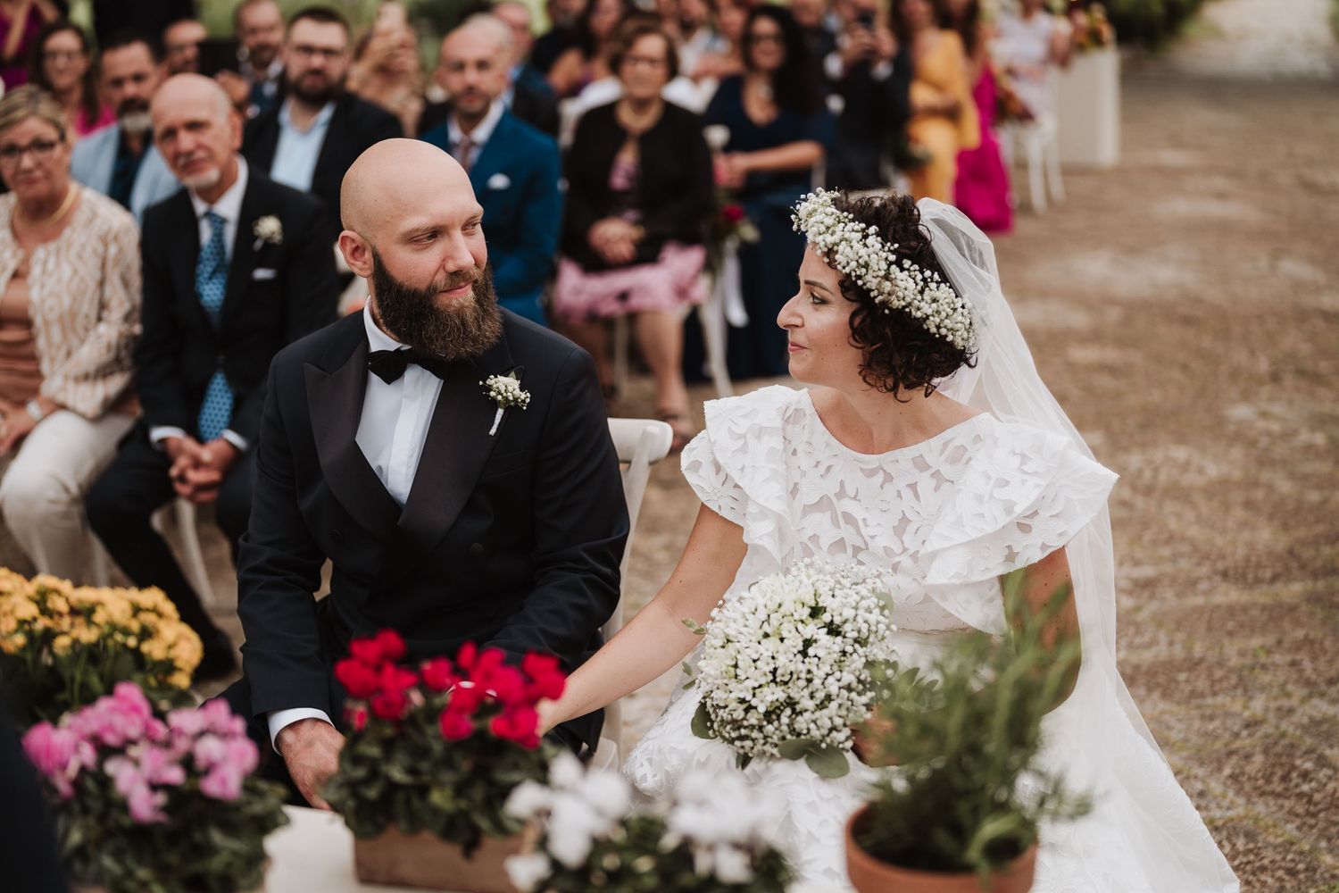 Elegant outdoor wedding ceremony with white floral decor and guests seated on white chairs.