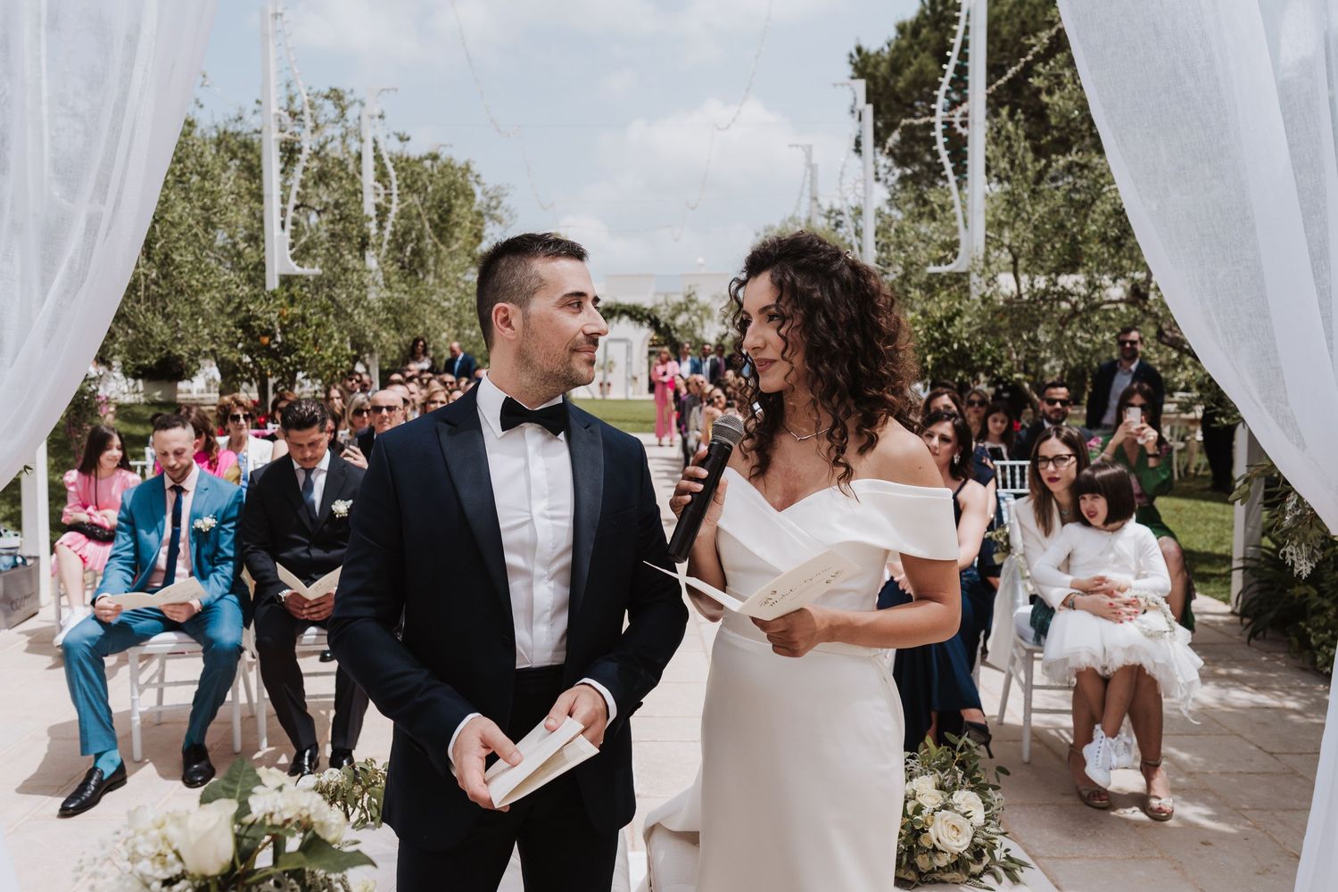 Couple exchanges vows under white draping during outdoor garden ceremony.
