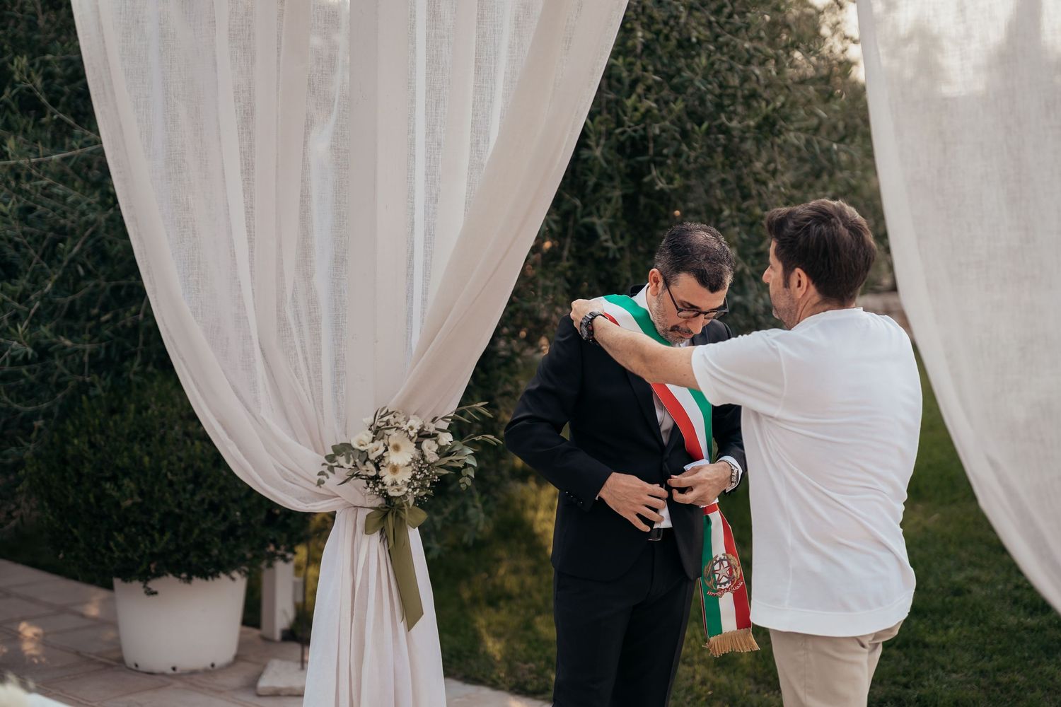Wedding ceremony moment with traditional tricolor sash exchange under white drapes outdoors.