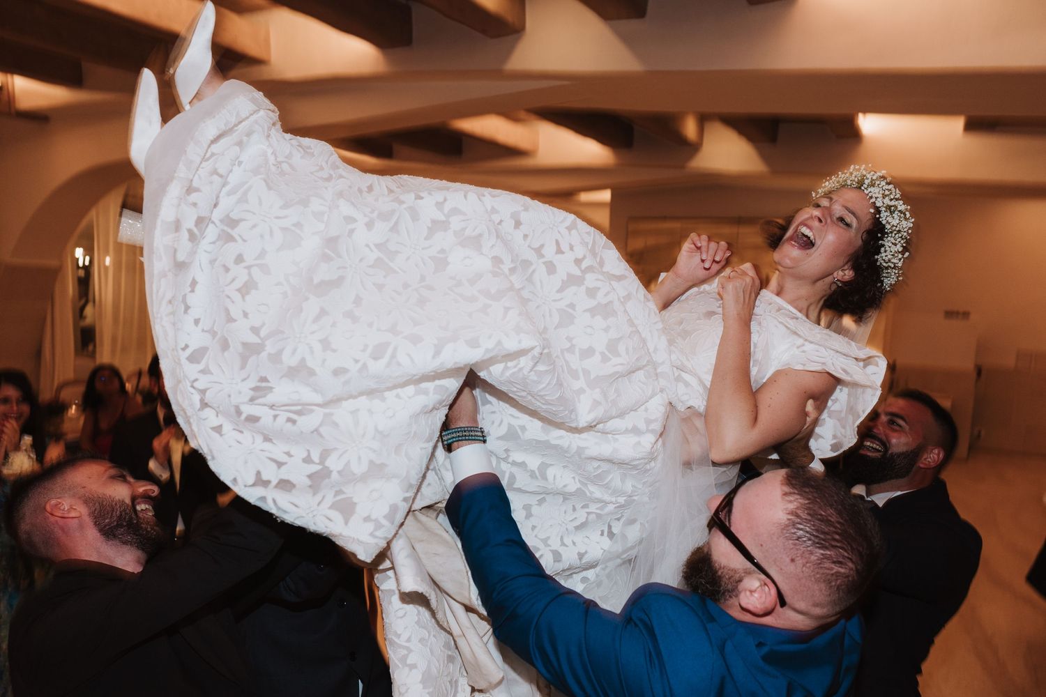 Wedding guests lift the bride in her lace dress during a joyful reception celebration.