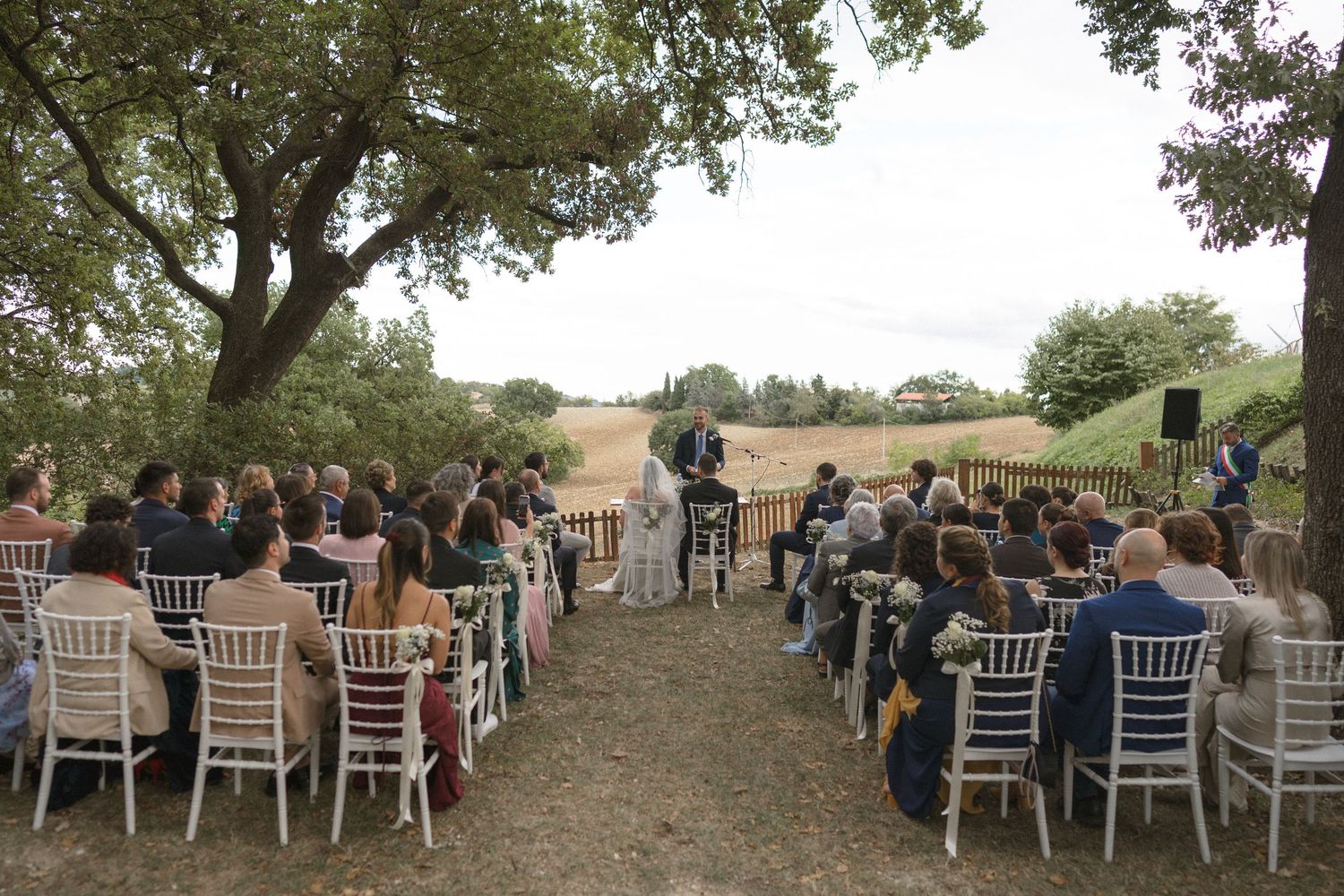 Outdoor wedding ceremony setup with white chairs arranged under large trees in a rustic setting.