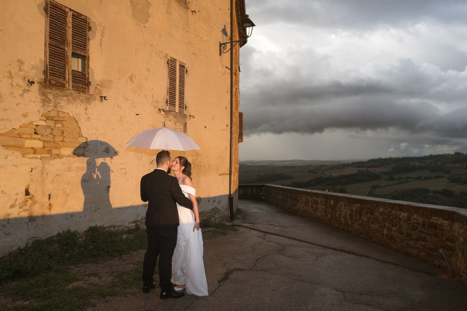 Dramatic cloudy sky over stone buildings with figures holding umbrellas silhouetted against warm walls.