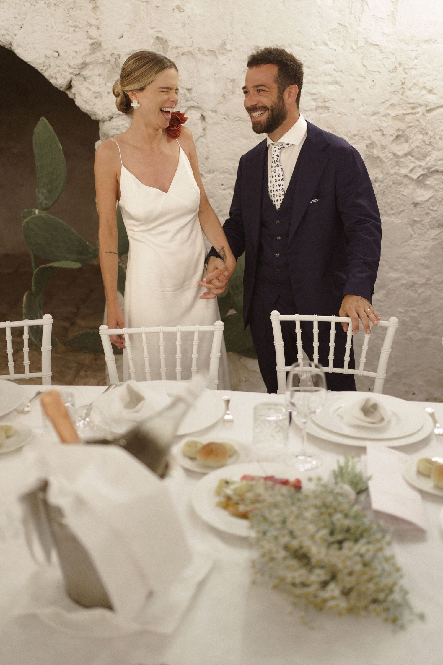 A joyful couple in wedding attire share a laugh while standing at an elegantly set reception table with white decor.