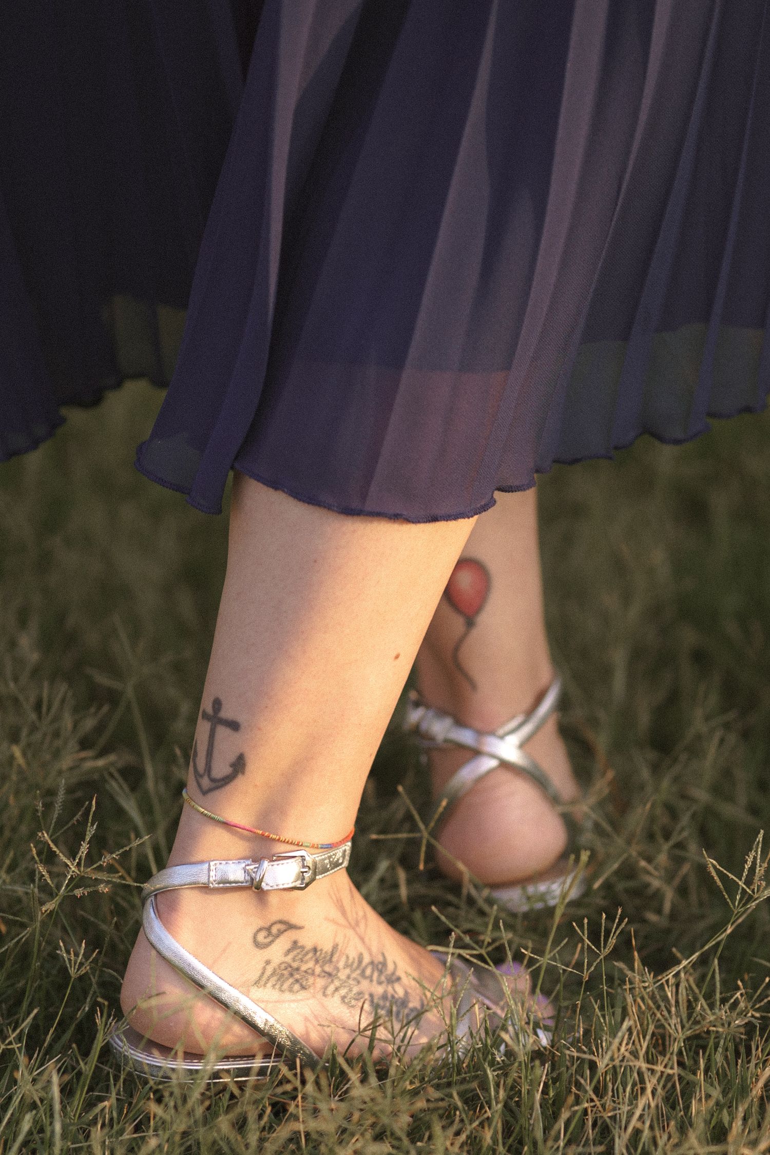 Close-up of silver sandals and anchor tattoo on ankle with blue dress hem and grass in background.