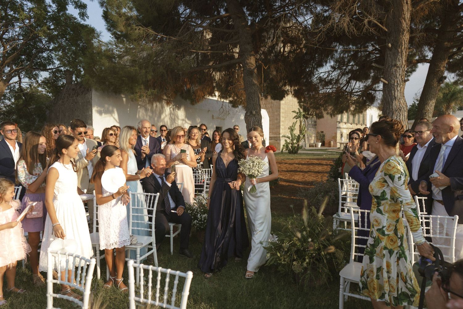 Outdoor wedding guests seated in white chairs gather around during a ceremony.