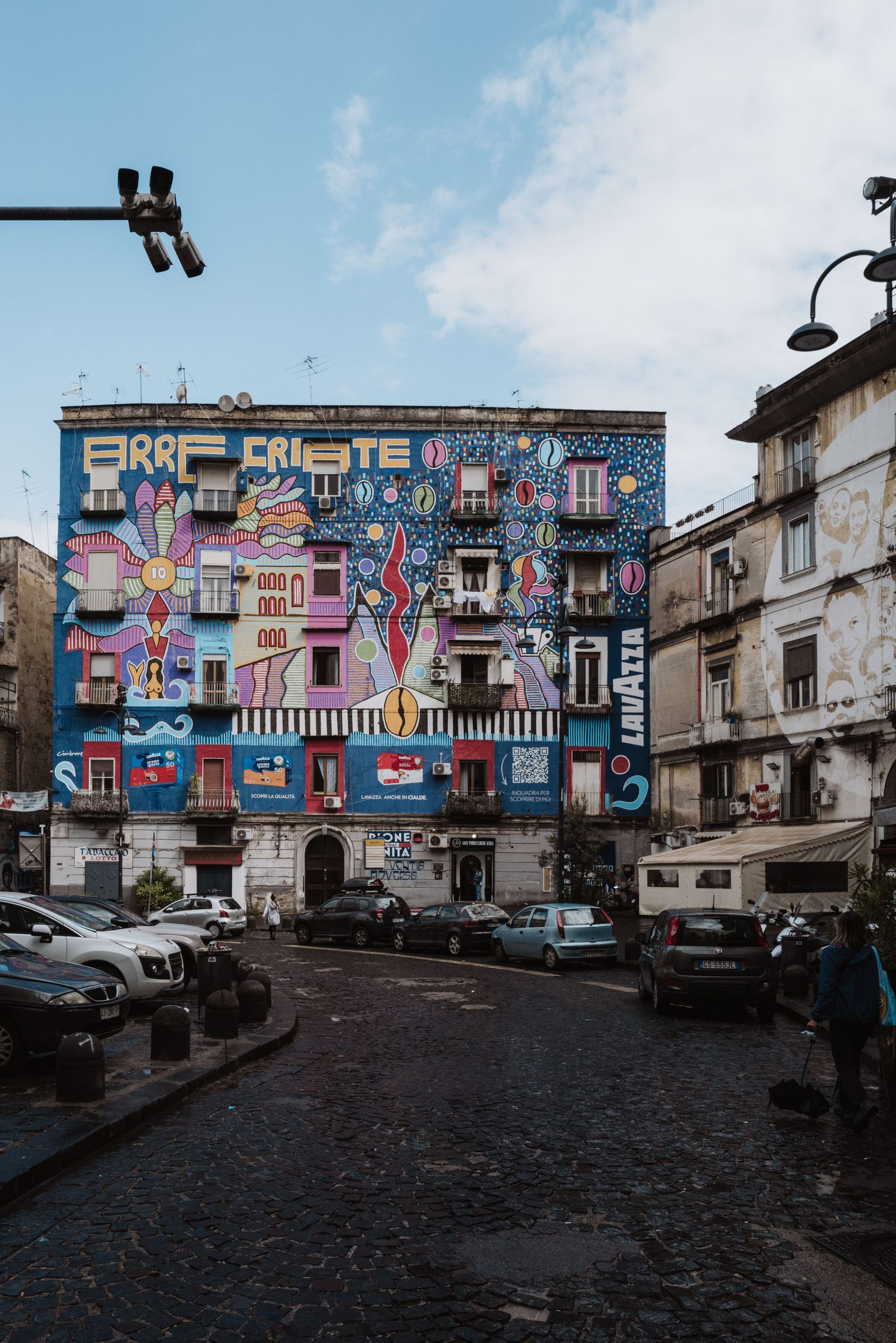 Colorful street art mural covers entire building facade in Naples with cars parked along cobblestone street.
