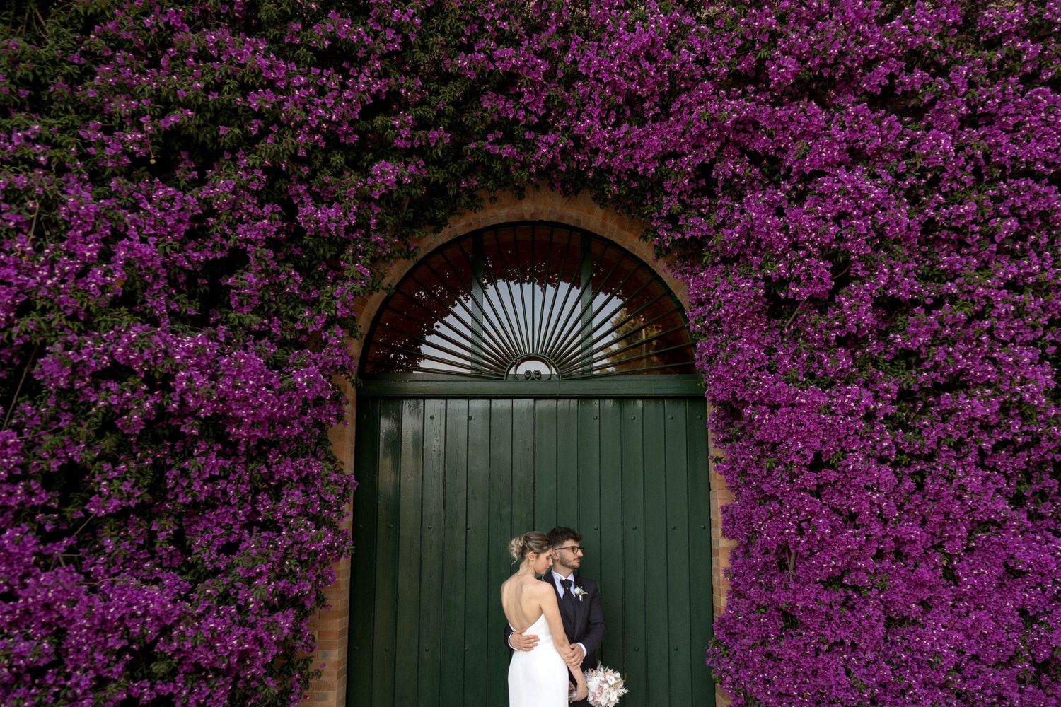 Vibrant purple bougainvillea flowers create a stunning natural arch over a series of green doorways along a stone wall.