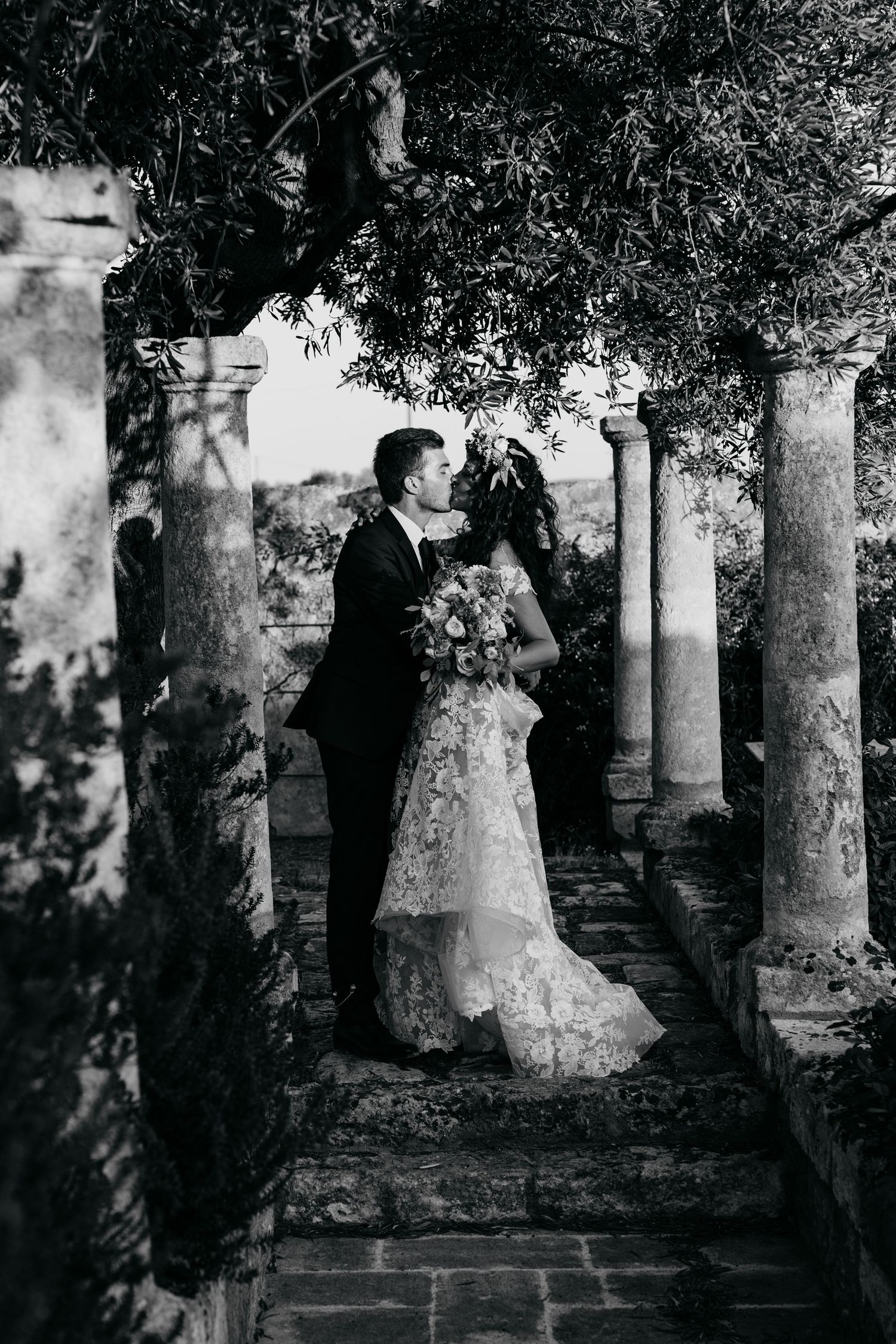 Romantic black and white wedding portrait among classical stone columns and greenery.