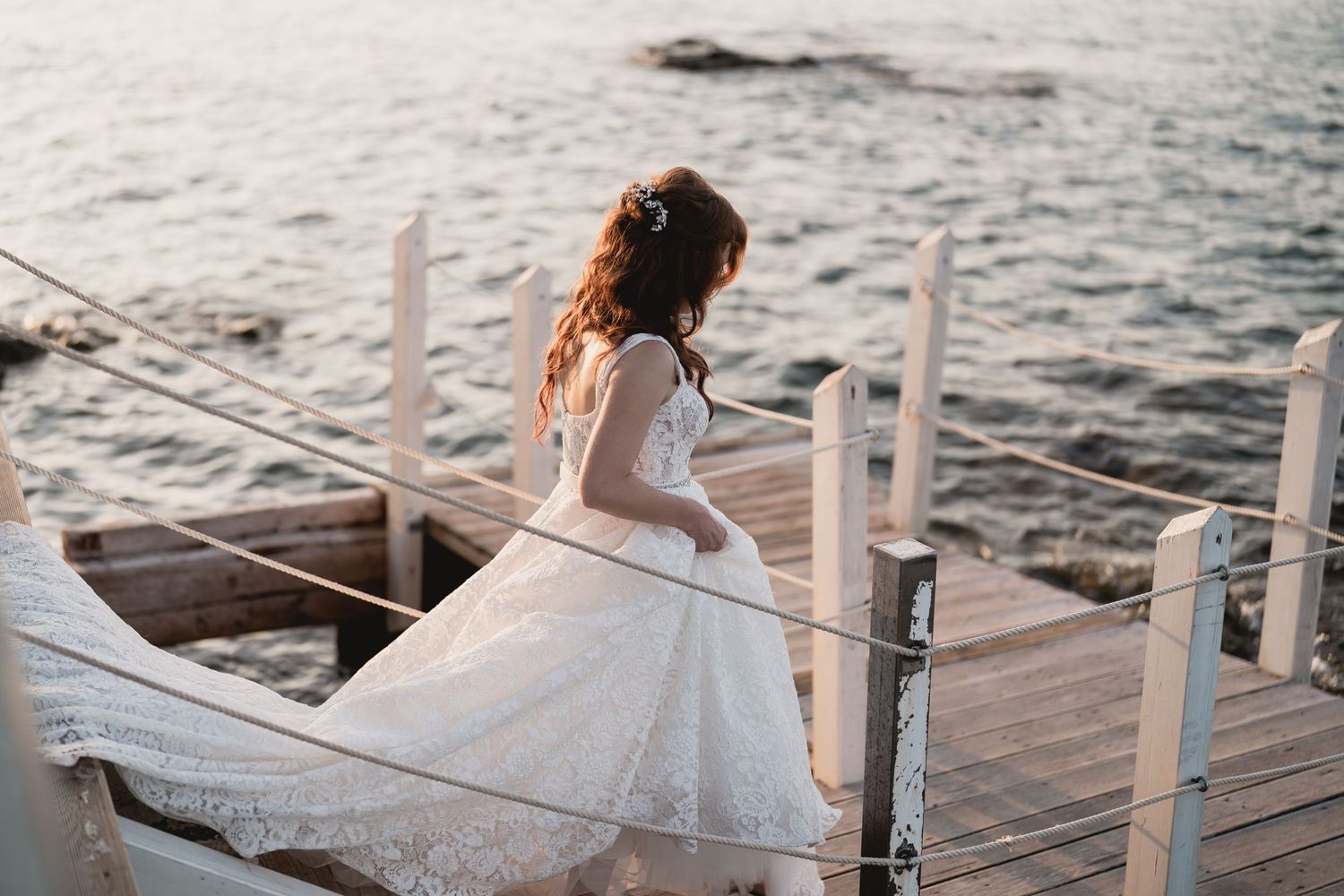 Bride in lace wedding dress poses gracefully on oceanfront dock at sunset.