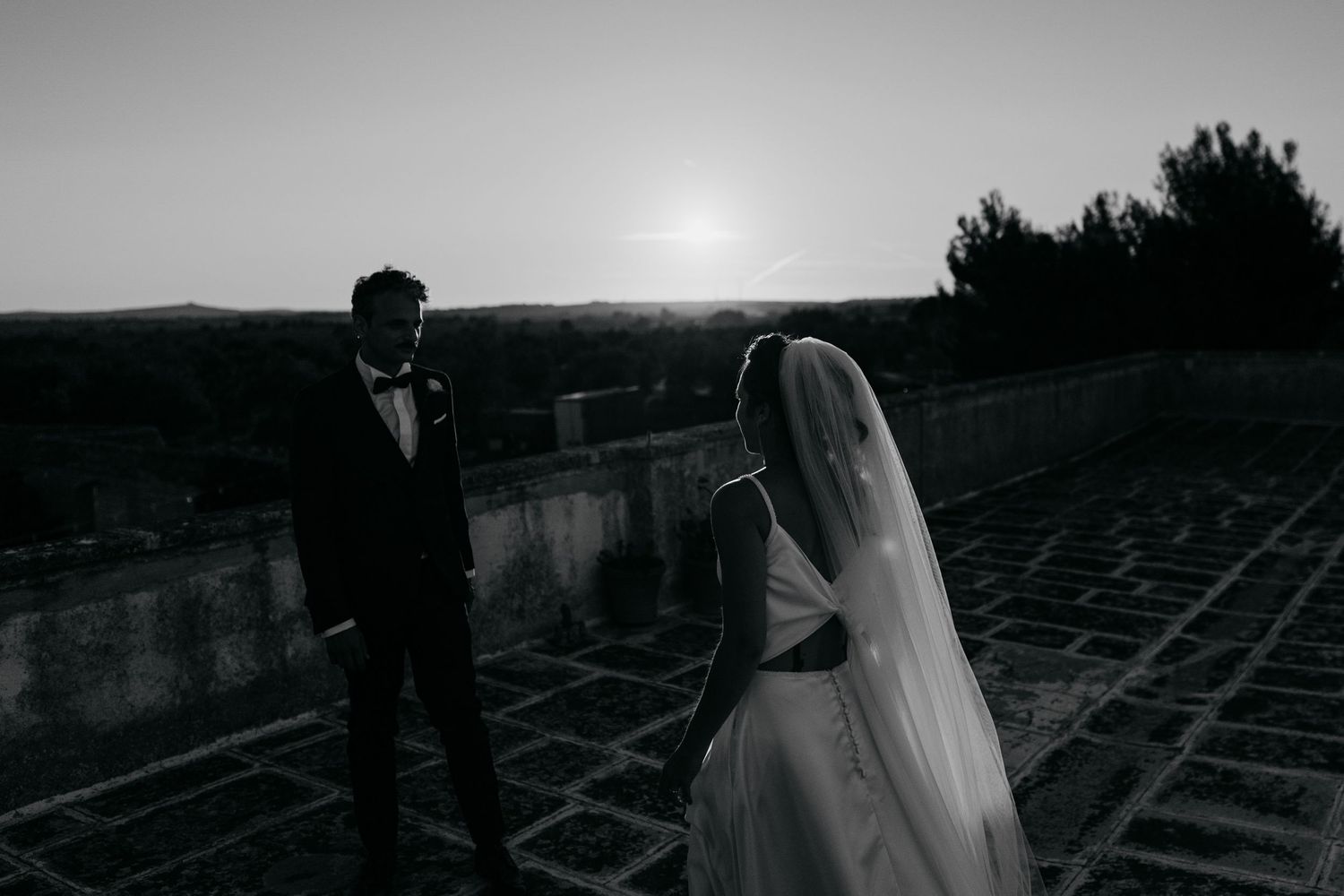 Black and white silhouette of newlyweds standing on a rooftop at sunset.