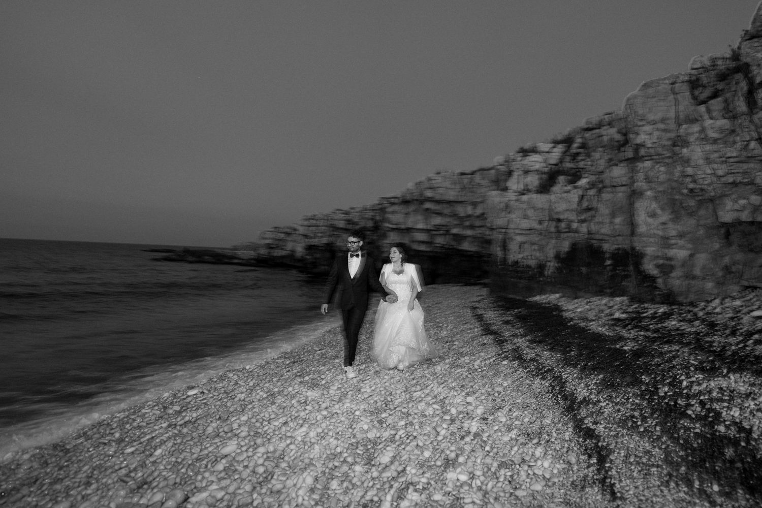 Black and white photo of a bride and groom walking hand in hand along a rocky beach with dramatic cliffs.