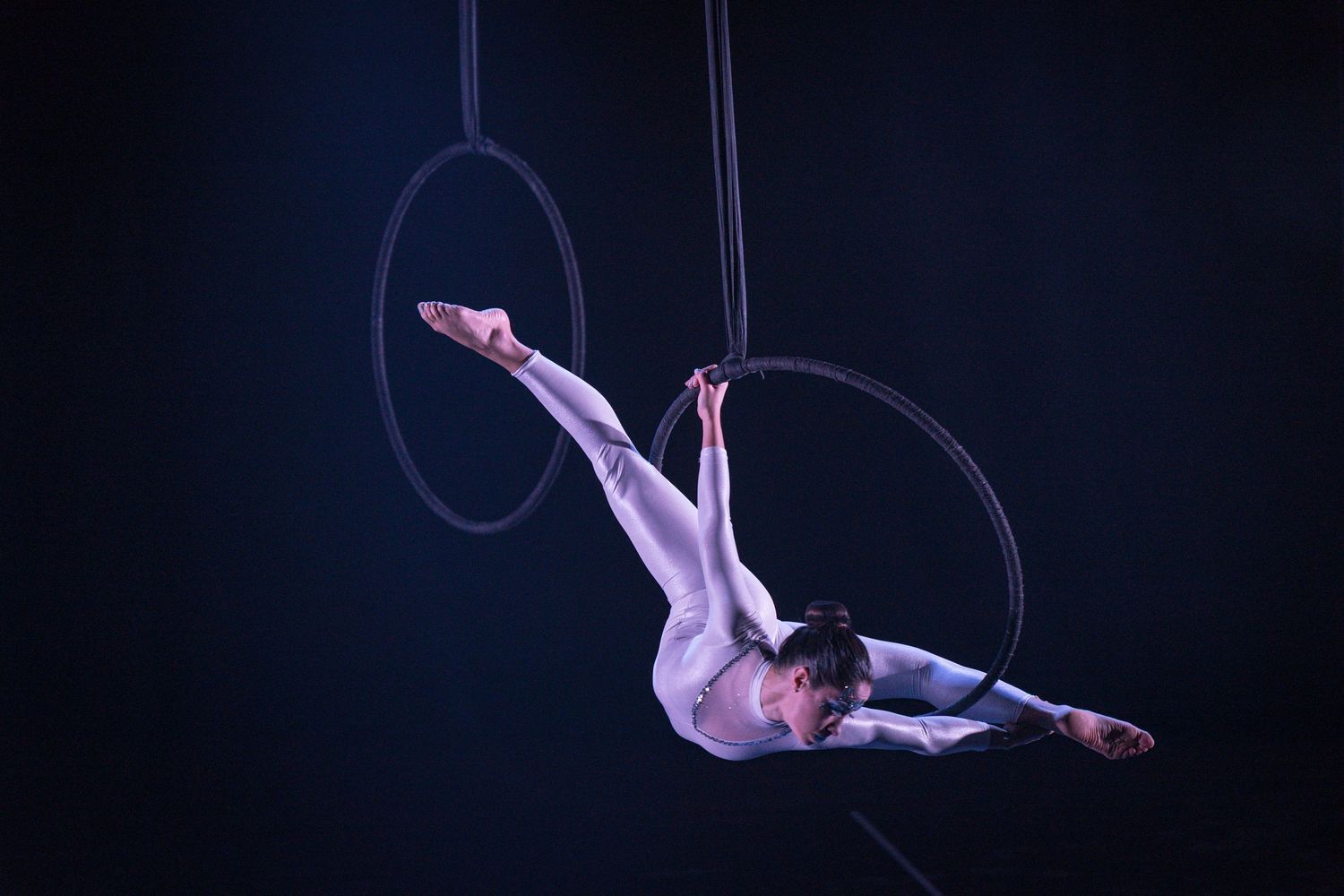 An aerial acrobat performs a graceful pose on suspended rings against a dark background during a circus performance.