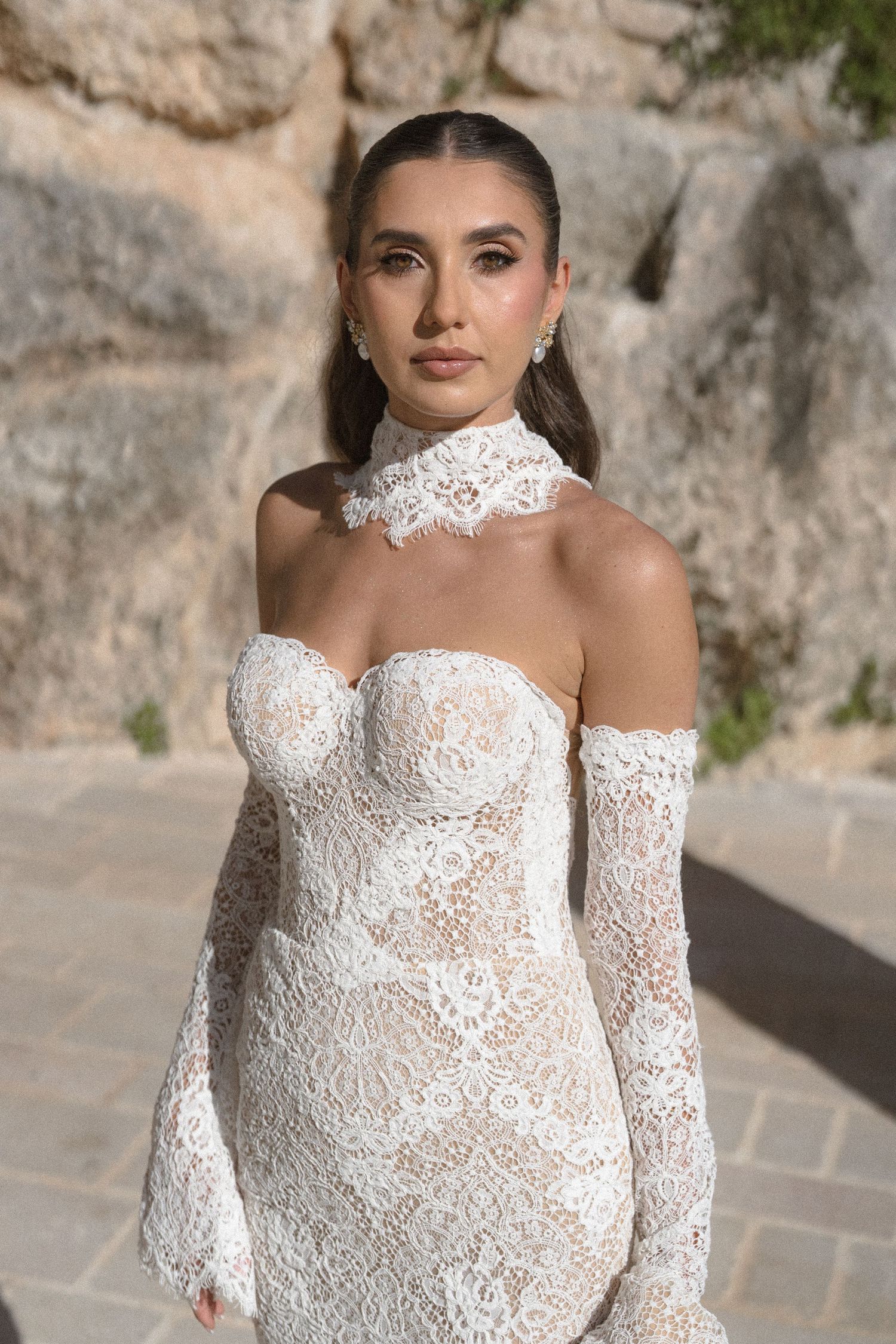 Bride in an off-shoulder white lace wedding dress with a lace choker collar and sleeves, posing before a stone wall.
