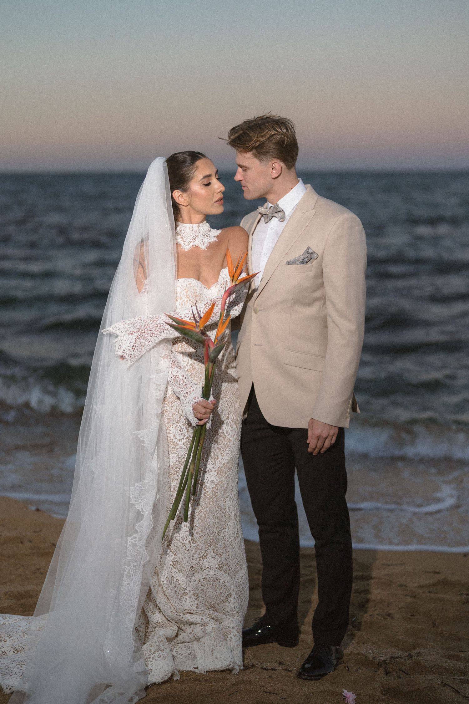 Bride in a lace wedding dress and veil with a bird of paradise bouquet embraces her groom on a beach at sunset by the sea.