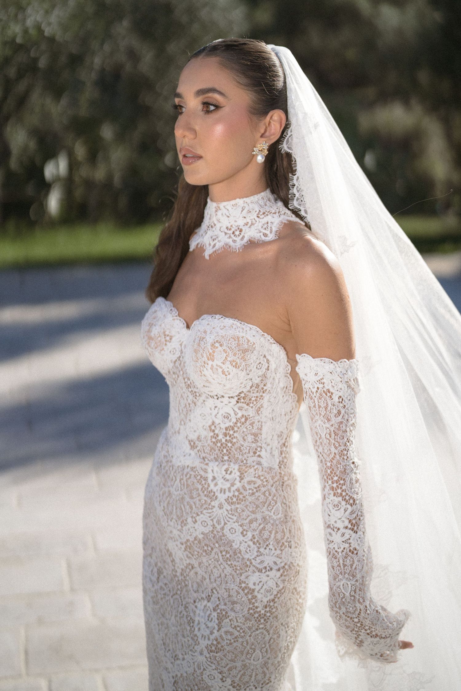 Bride in a strapless lace wedding gown with a lace collar, long sleeves and a flowing veil standing outdoors.