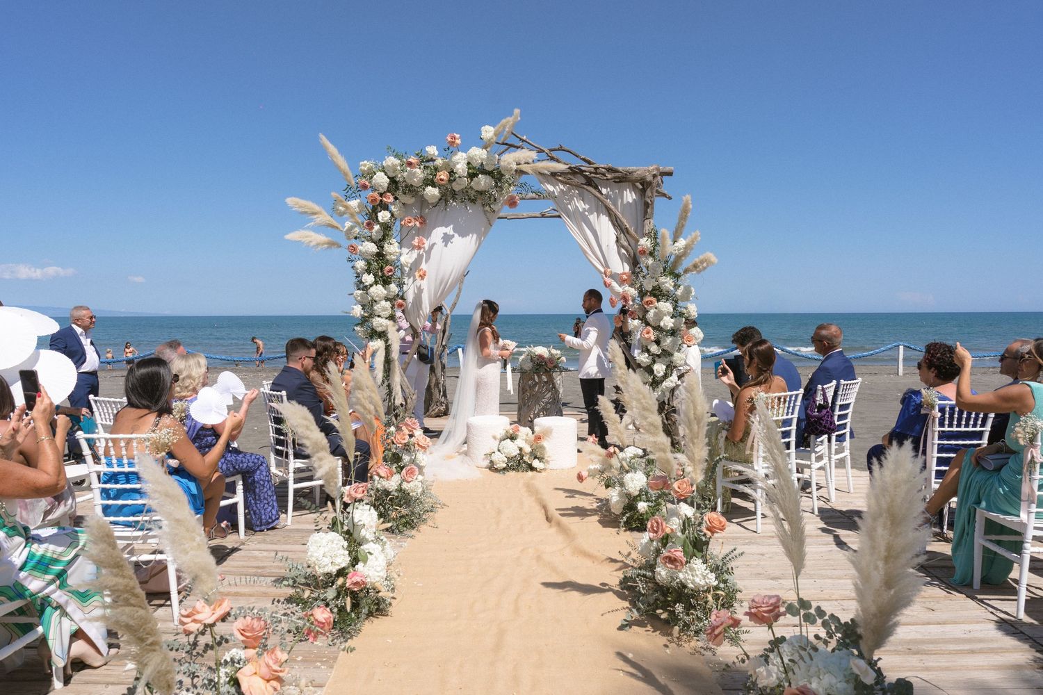 Beach wedding ceremony setup with floral arch against blue ocean backdrop.