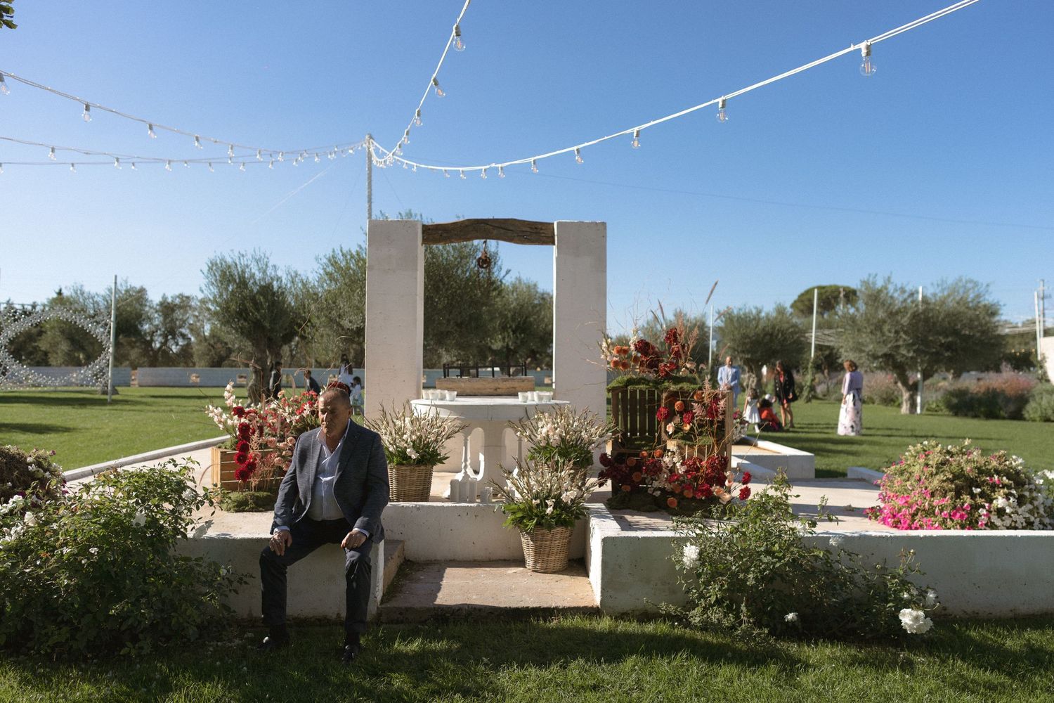 Man sitting beside an elegant outdoor wedding altar decorated with flowers and string lights in a sunny garden.