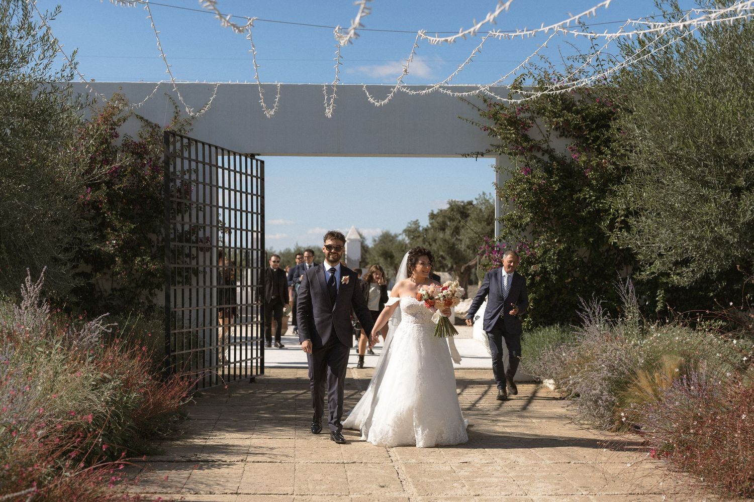 Bride and groom walk through decorated garden gate with wedding guests at outdoor venue.