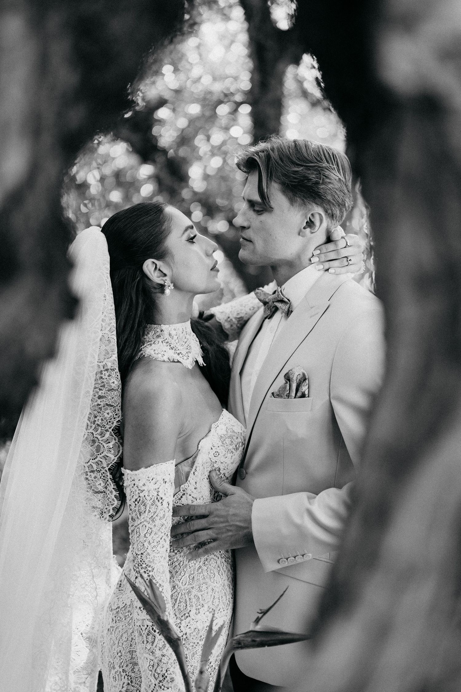 Black and white photo of a bride in a lace gown and veil embracing her groom in a blazer, framed by blurred tree branches.