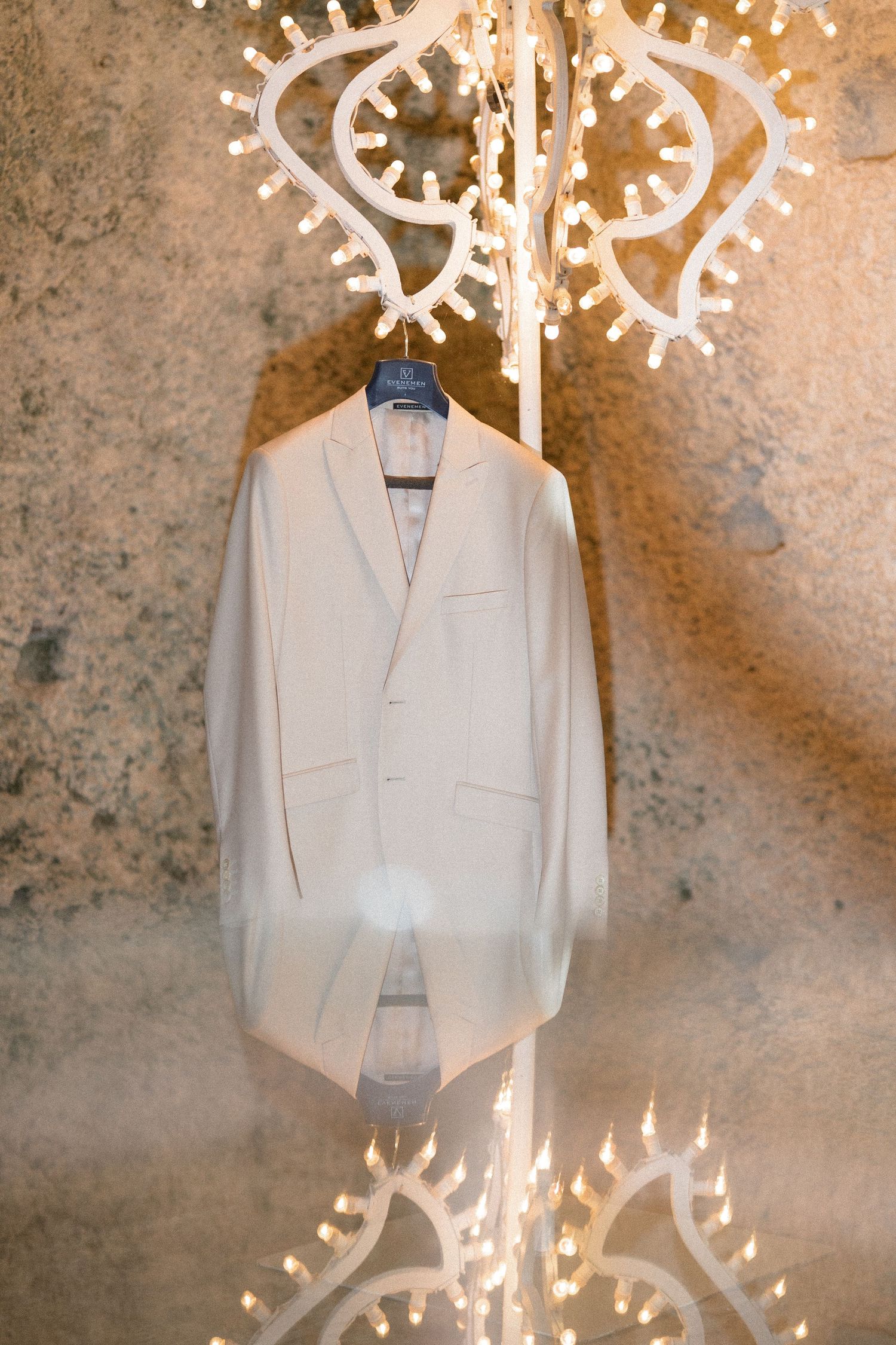 Cream wedding suit jacket hanging on a dark hanger against a rustic stone wall, framed by ornate decorative string lights.