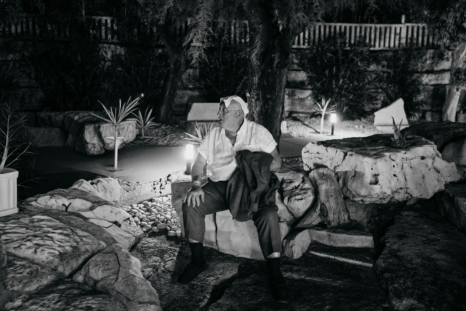A man sits on rocks by candlelight in a dark garden at night in a black and white photo.