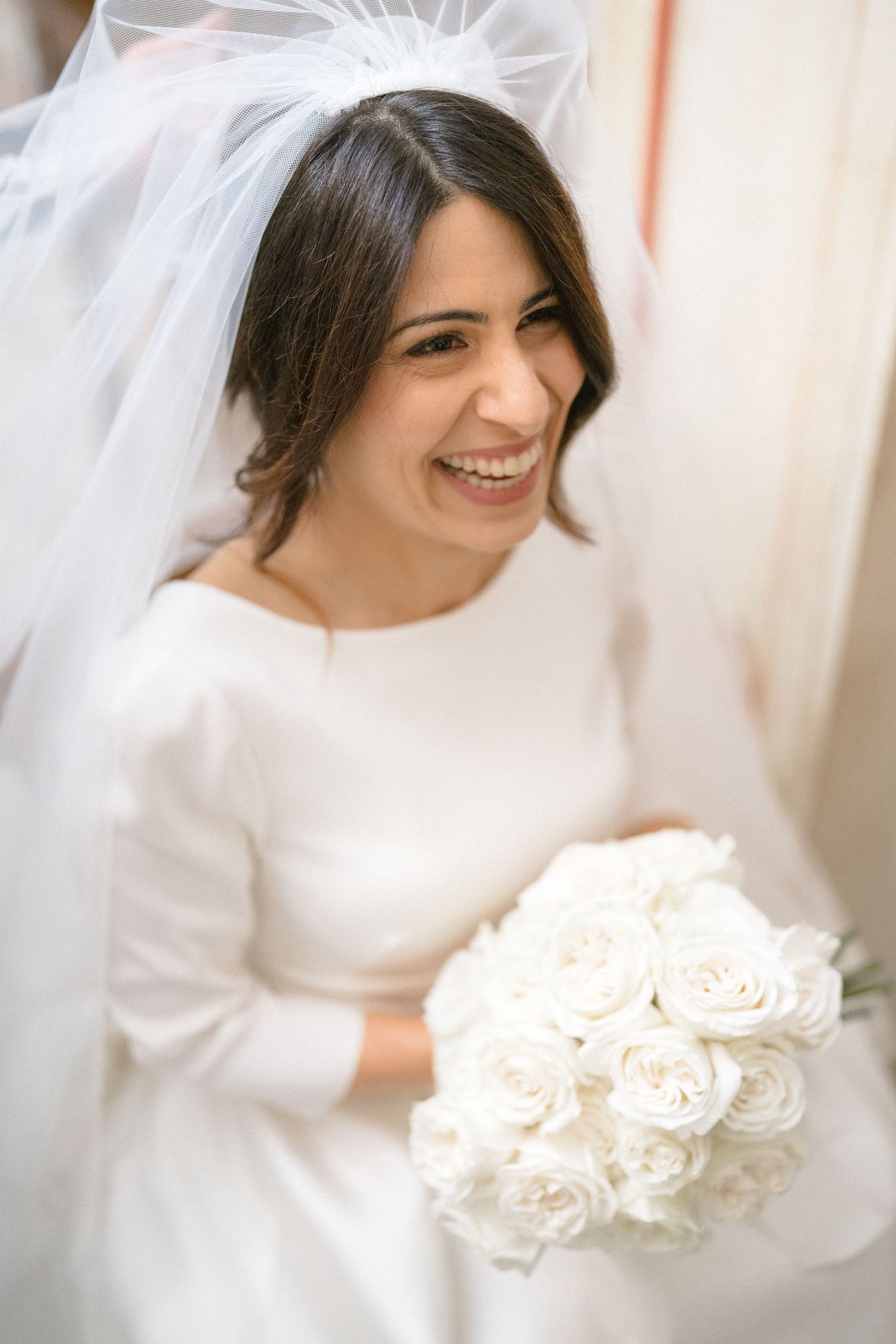 Smiling bride in white gown and veil holding a bouquet of white roses on her wedding day.