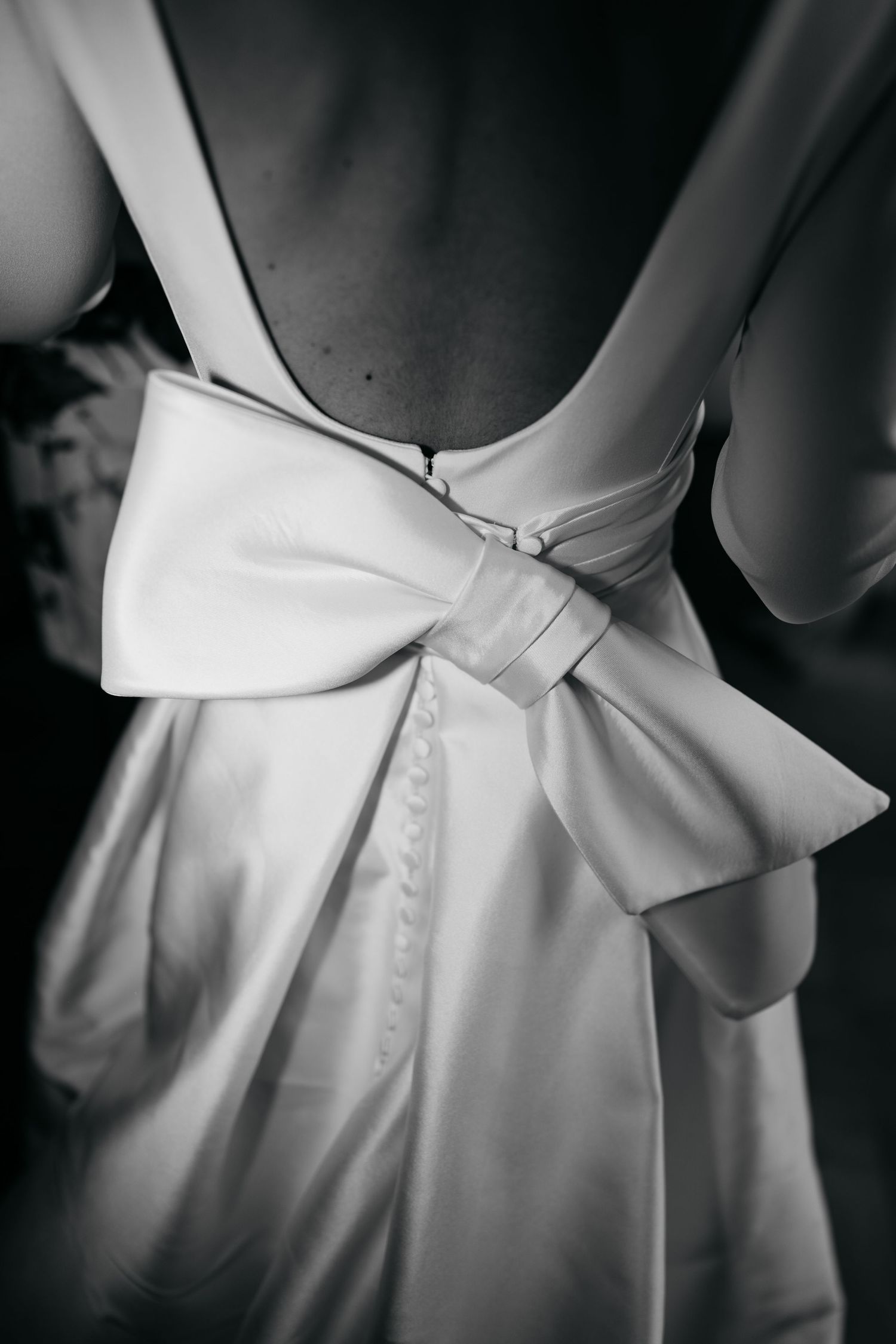 Black and white close-up of elegant backless wedding dress featuring large satin bow and button detail.