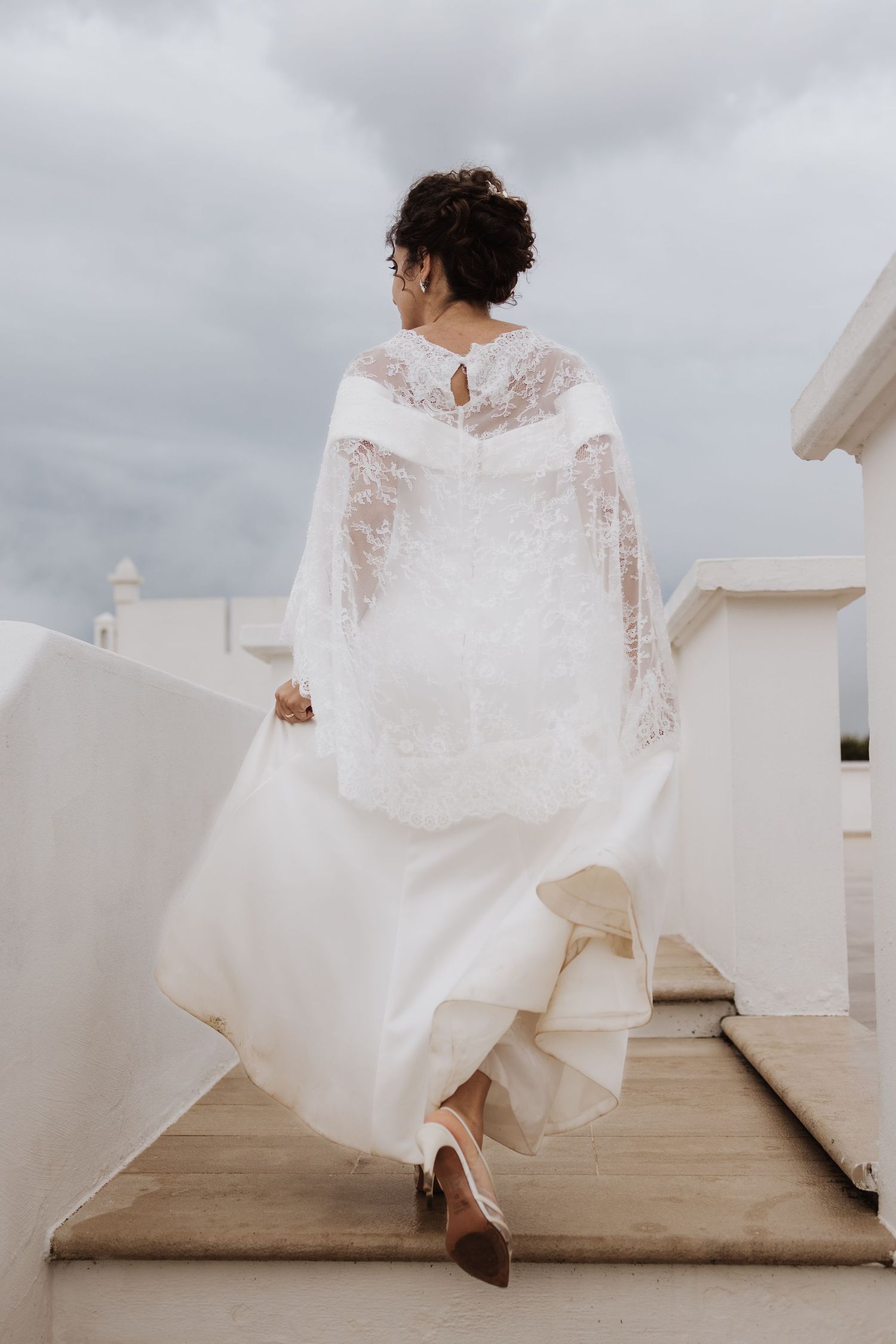 Back view of an elegant white lace wedding dress with flowing fabric on an outdoor terrace.
